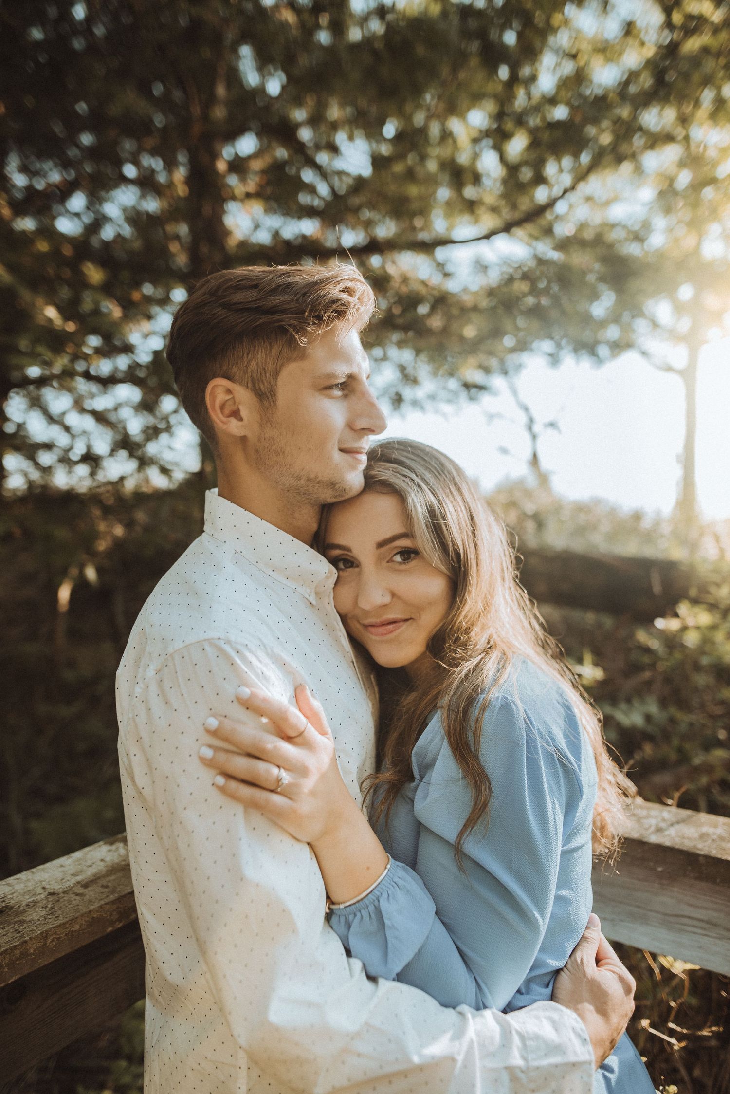 A romantic couple embraces in golden sunlight among trees, wearing white and blue casual attire.