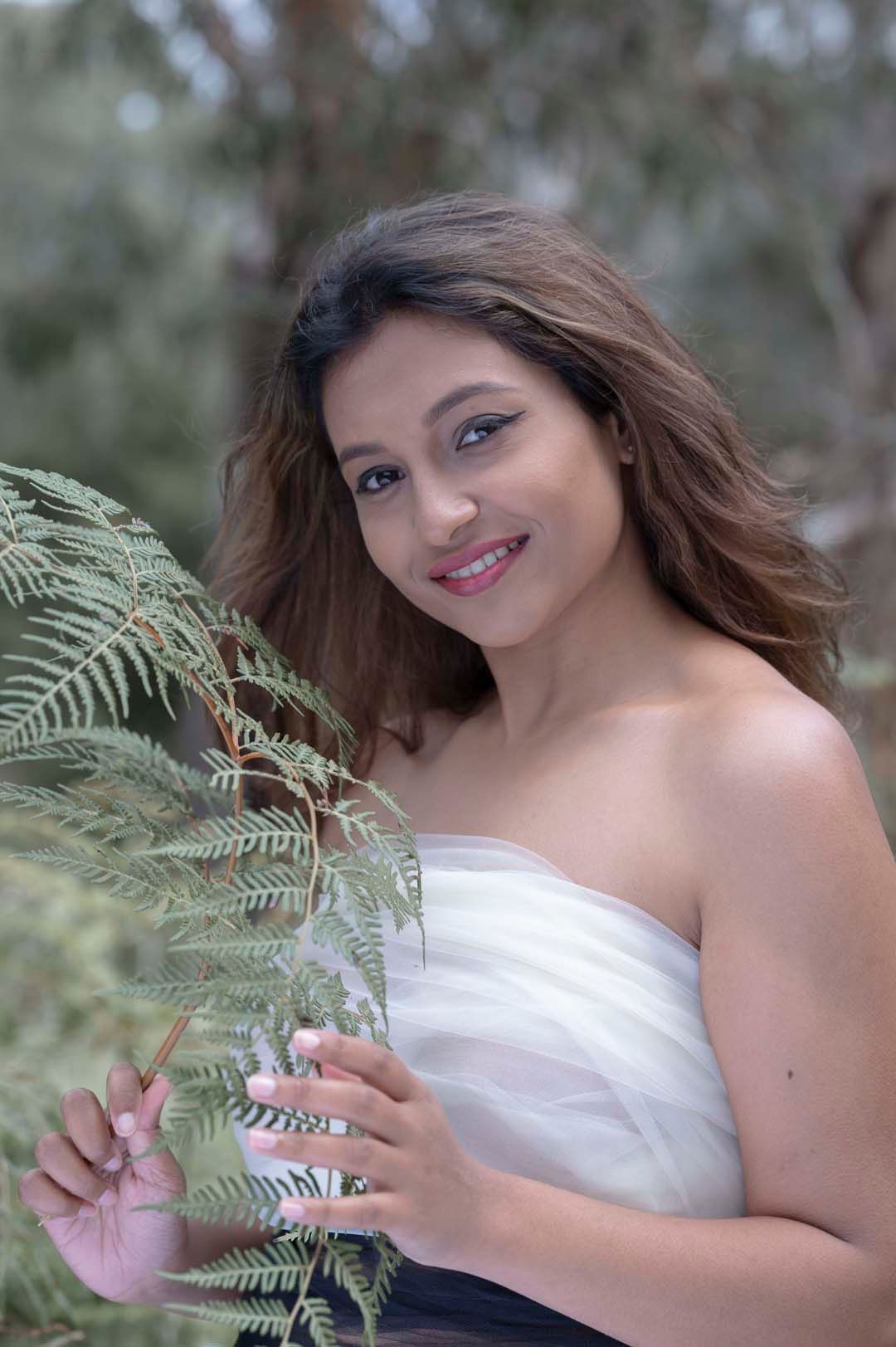 Beautiful smiling woman in white strapless top posing beside green fern leaves in a lush outdoor setting.