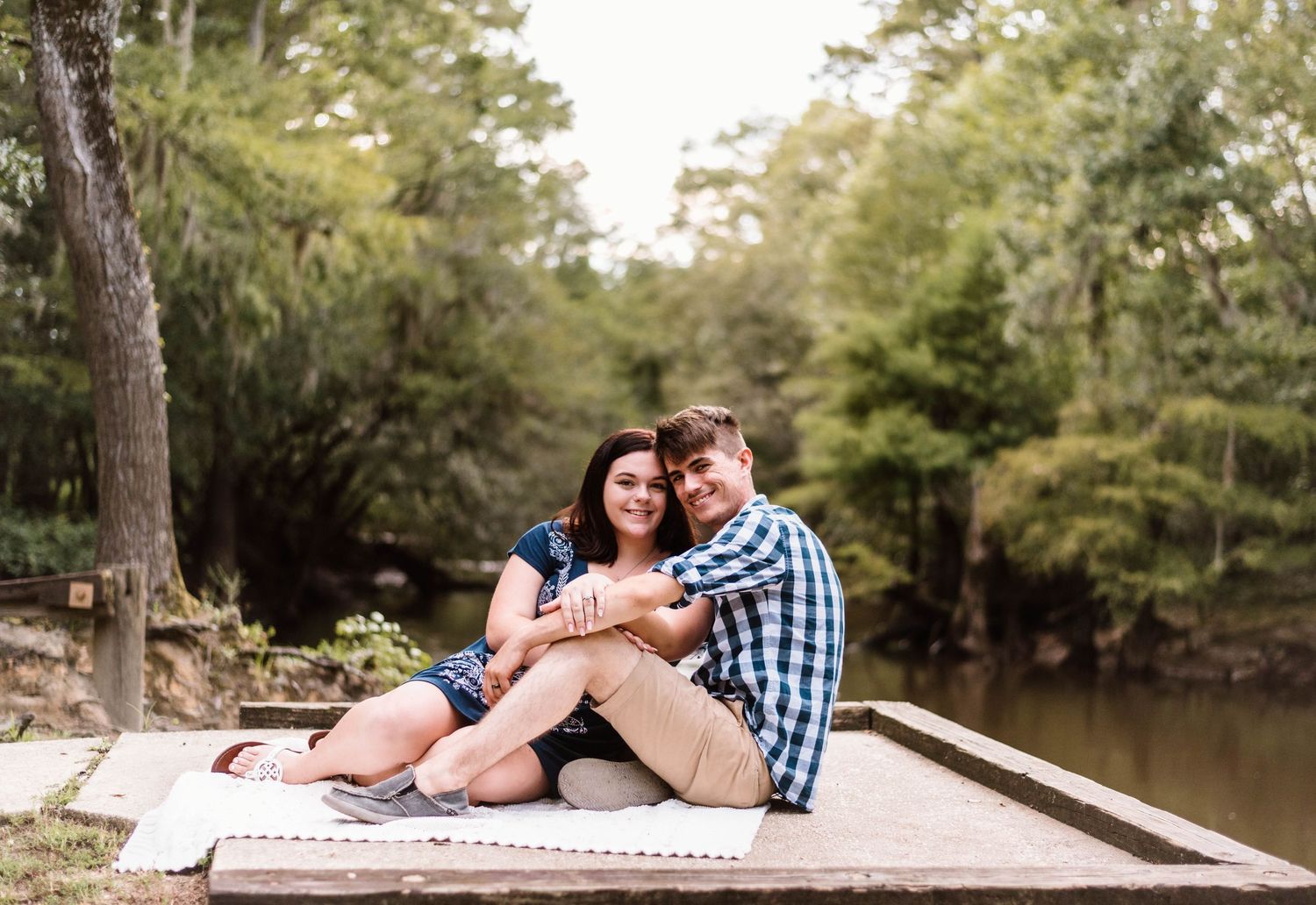 Romantic couple lounging by a peaceful lake surrounded by pine trees on a sunny afternoon.