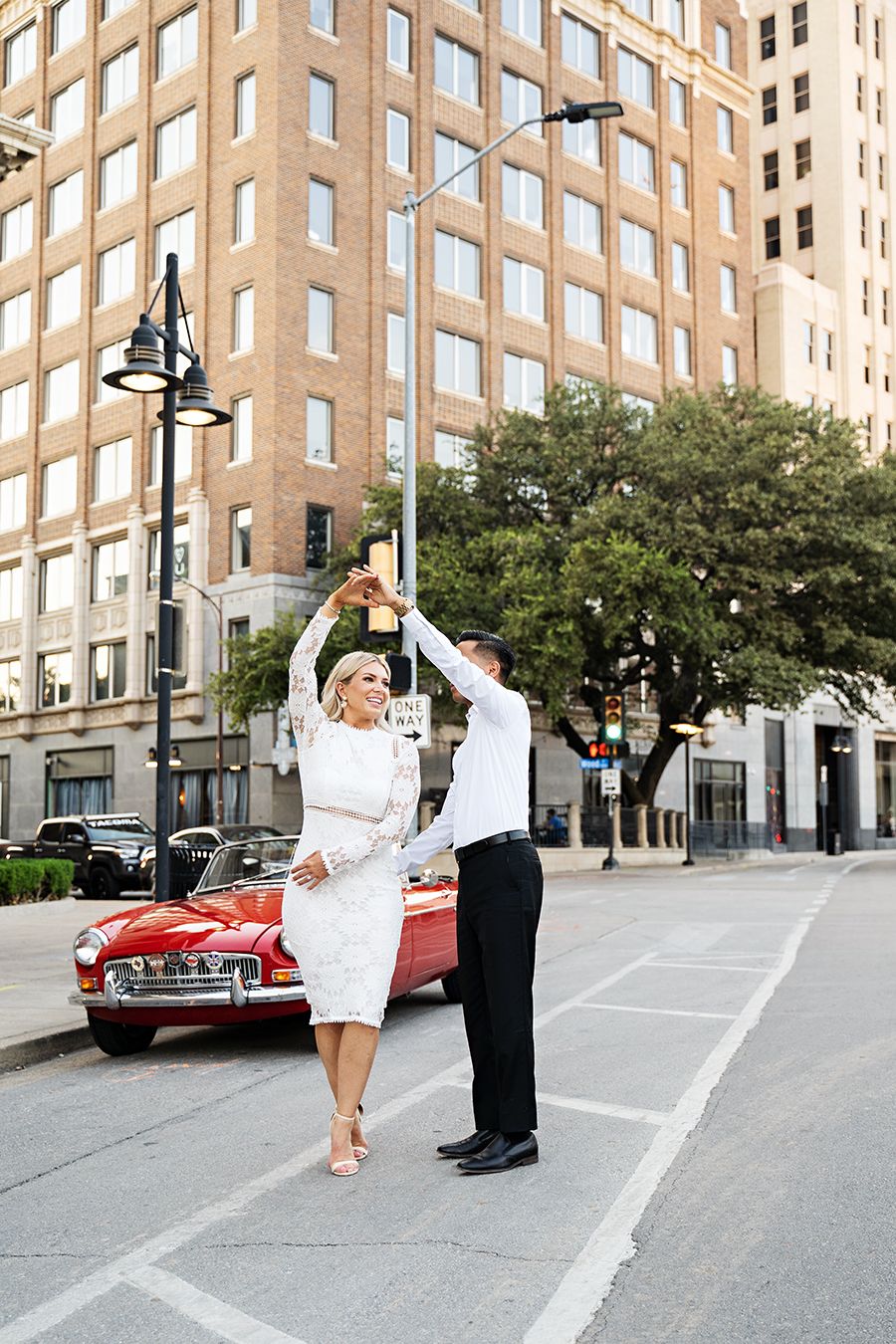 Dancing couple photographed against urban architecture with a red vintage car.
