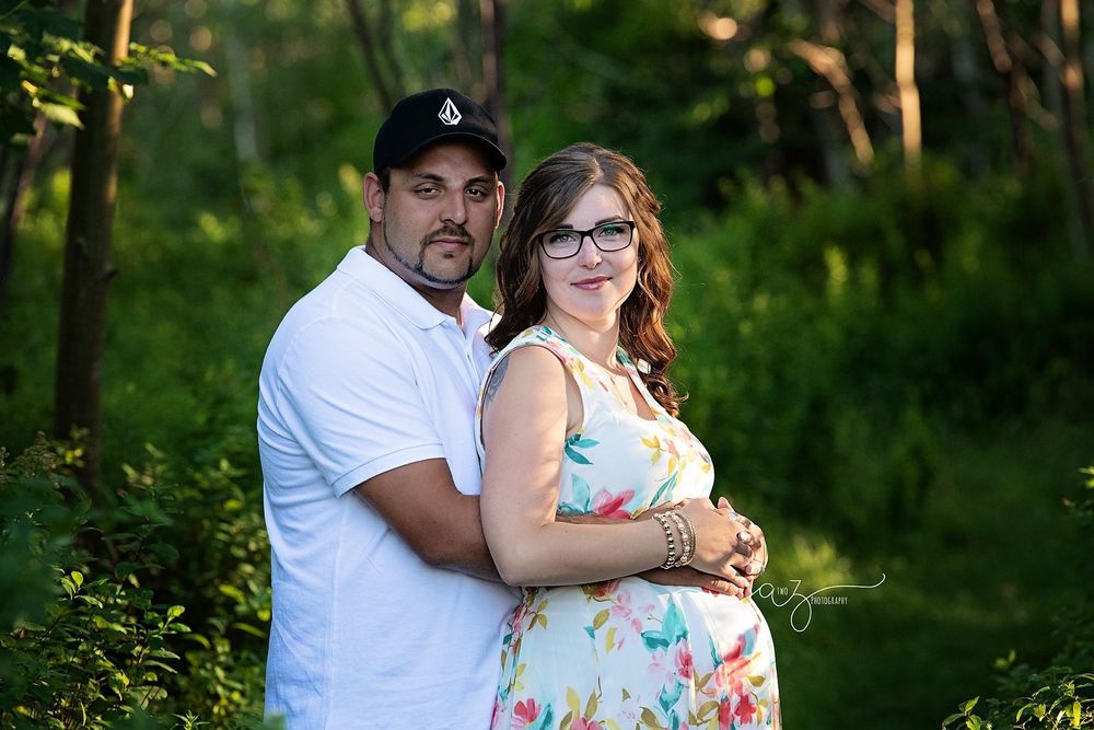 An expectant couple poses together against a backdrop of trees in summer.