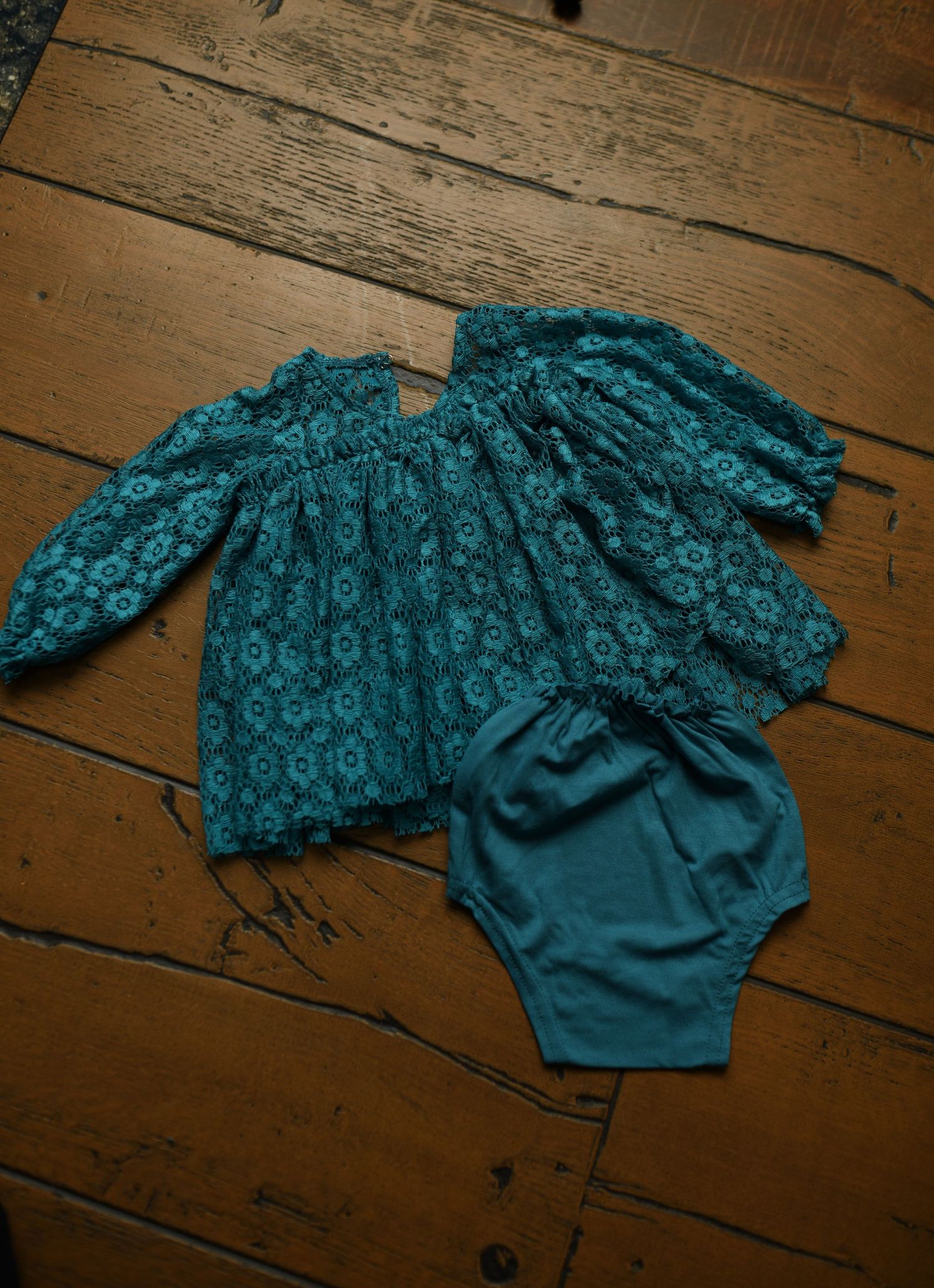 Teal floral patterned baby romper displayed on rustic wooden floor.