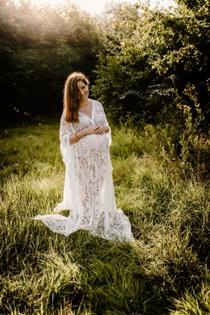 Ethereal maternity portraits in flowing white lace dress amid tall summer grass at golden hour.
