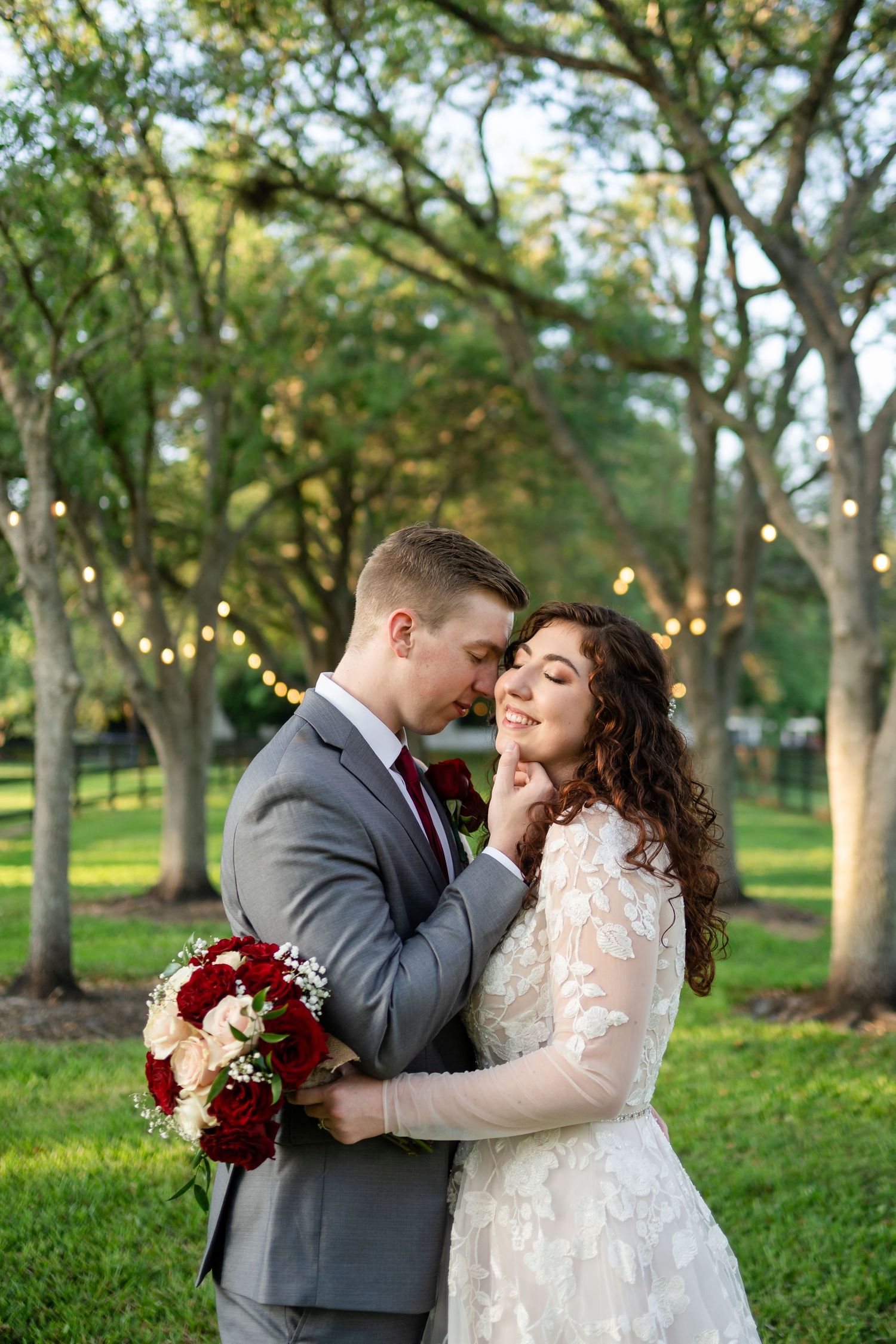 Romantic outdoor wedding portrait with couple embracing under string lights and trees.