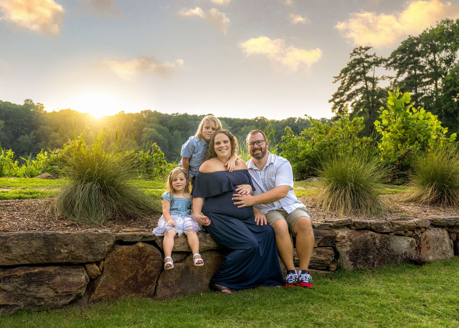 Family sits together on stone wall during golden sunset in landscaped garden setting.