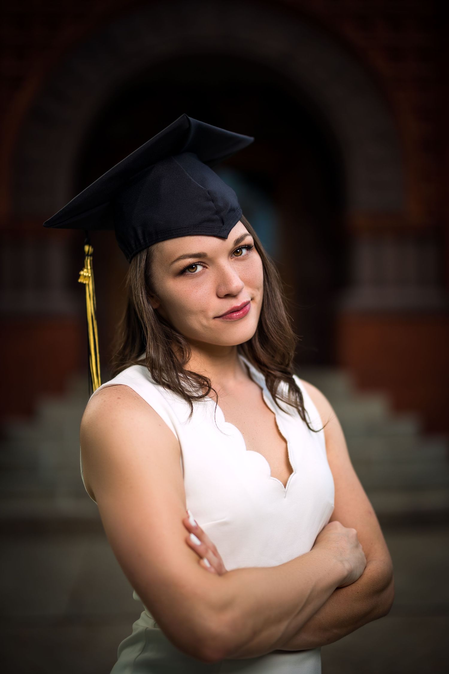 Graduate in white dress and cap poses confidently against dark architectural background.