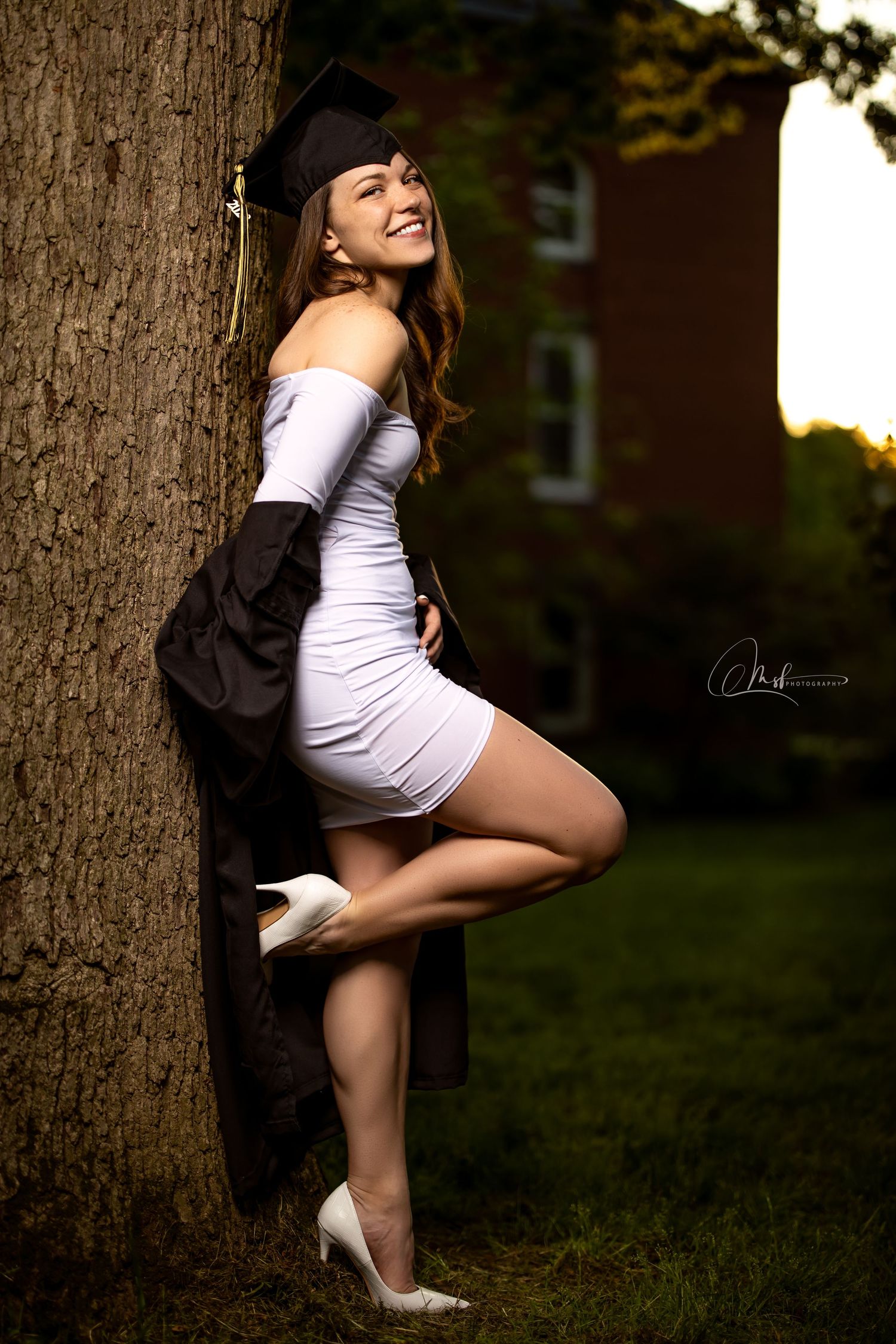 Playful graduation photo with cap and gown against tree at sunset.
