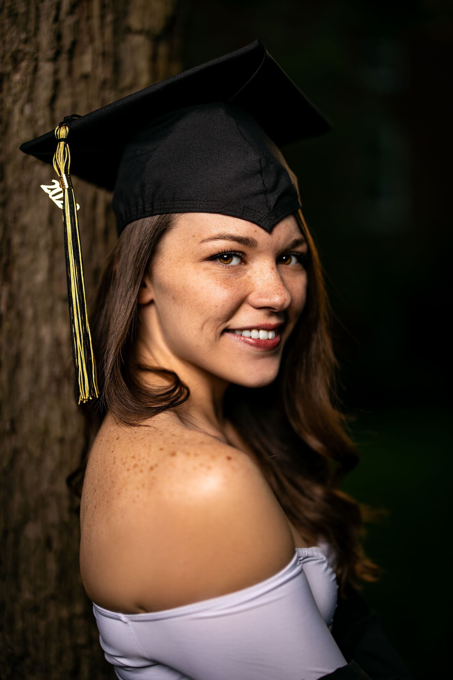 Graduate in black cap with tassel poses against tree trunk in natural lighting.