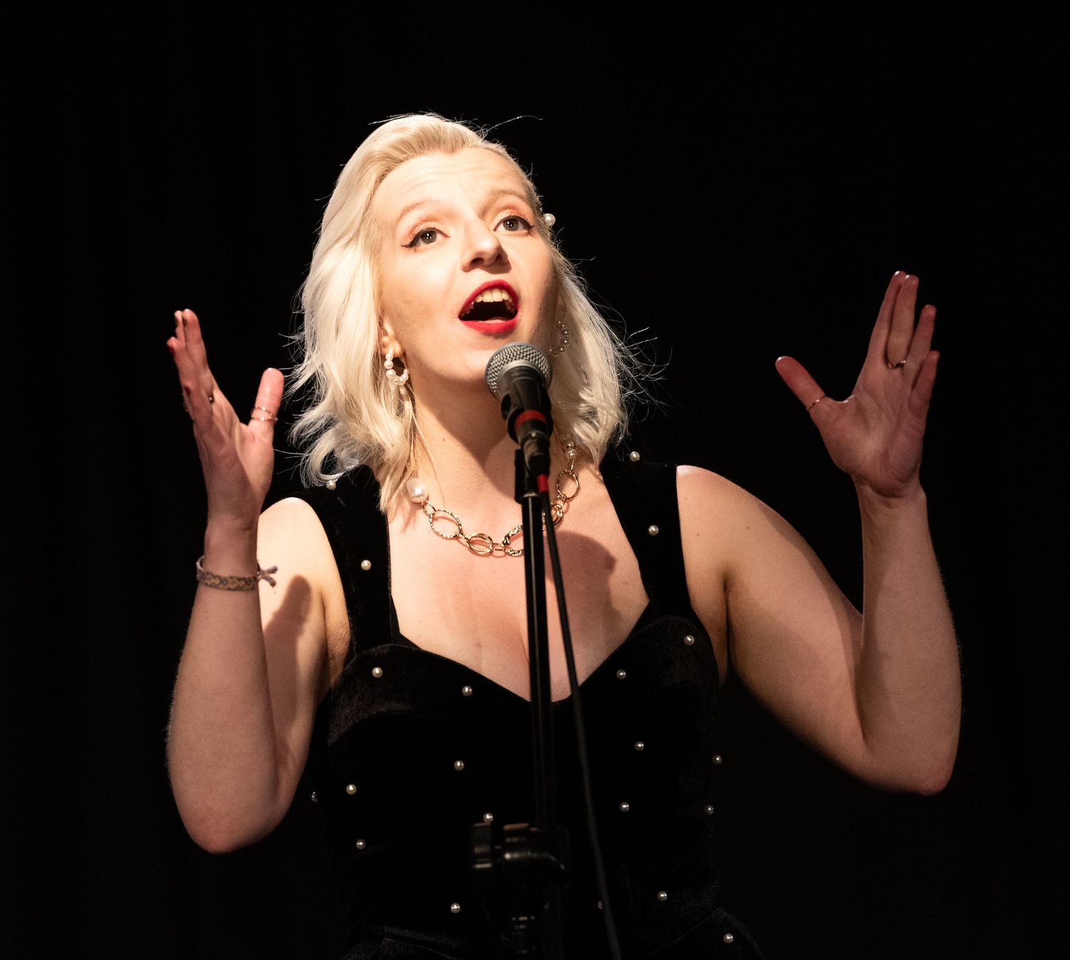 A performer in a black sequined dress sings emotionally on stage with raised hands.