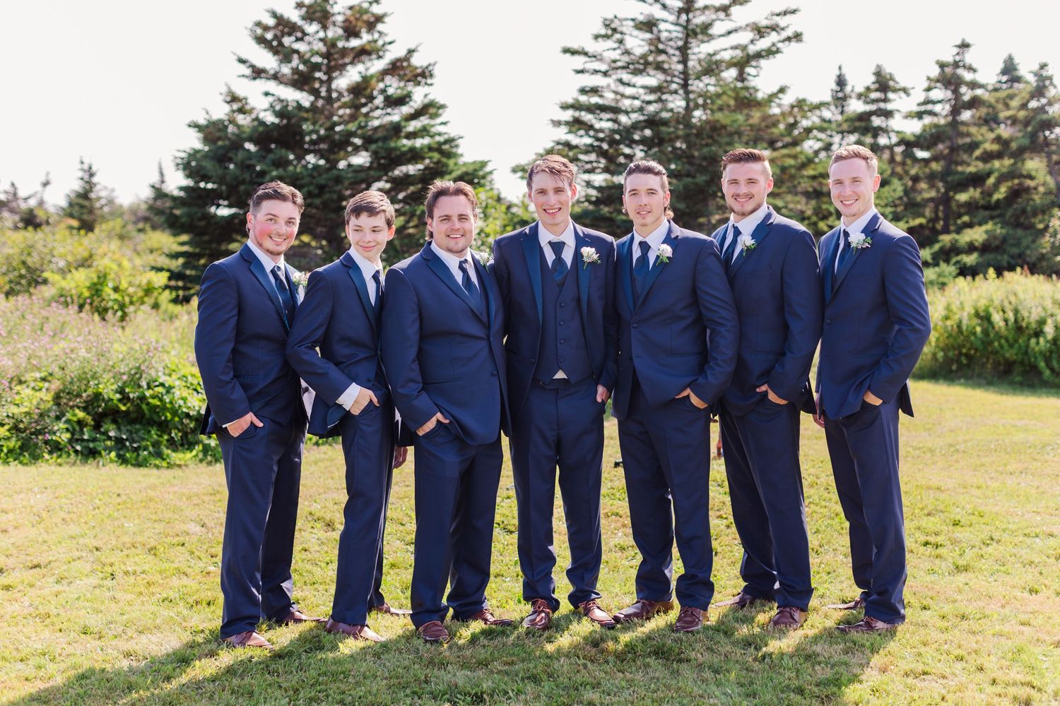 Wedding party groomsmen in navy suits stand together in a line outdoors with evergreen trees in background.