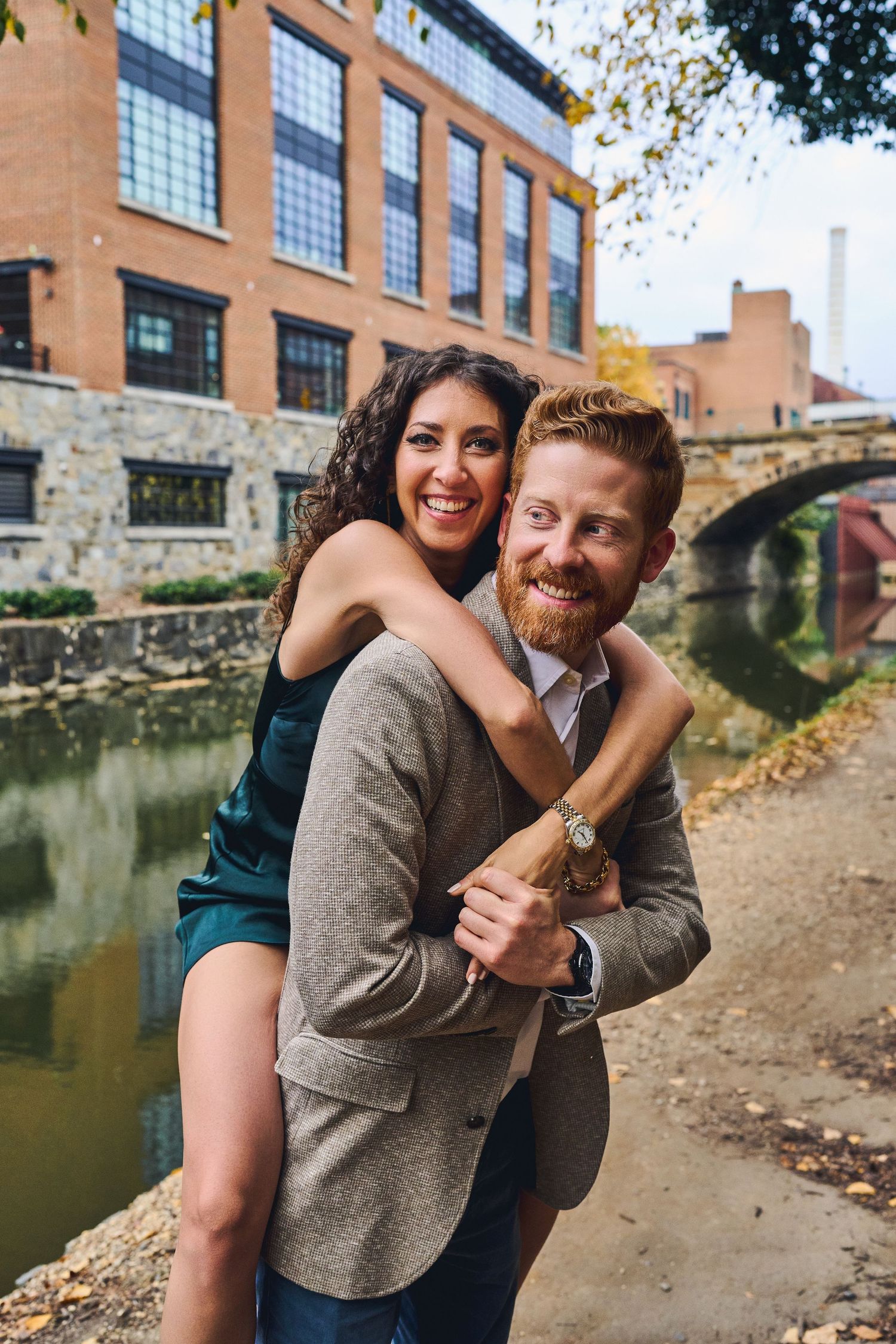Playful couple posing by canal with brick buildings and autumn trees in background.