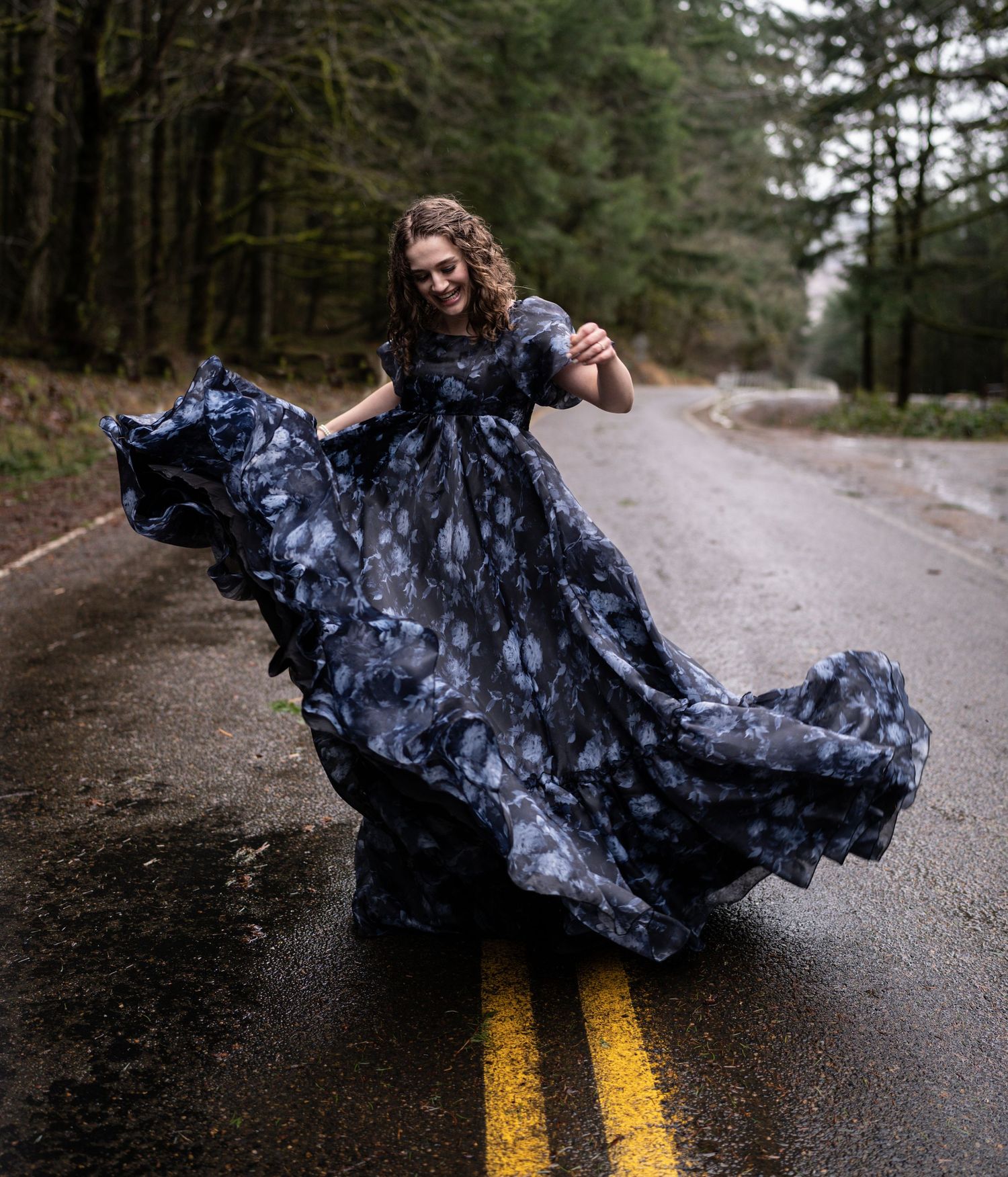 A person twirling in a flowing blue dress on a rainy forest road.