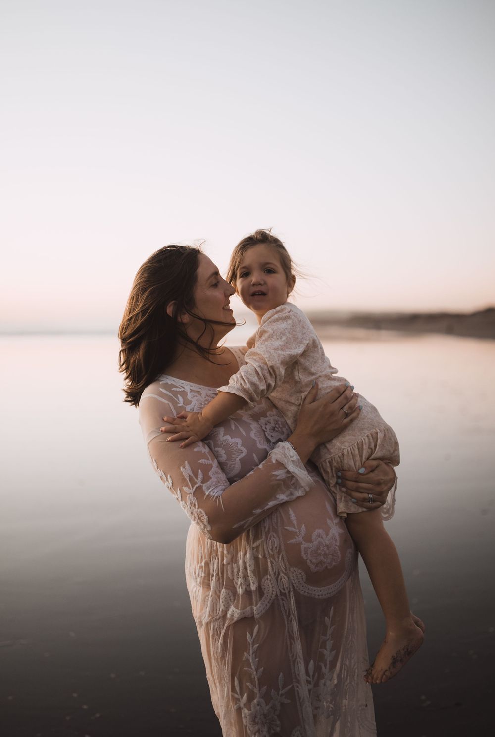 Mother and child embracing on beach during sunset in white lace dress.