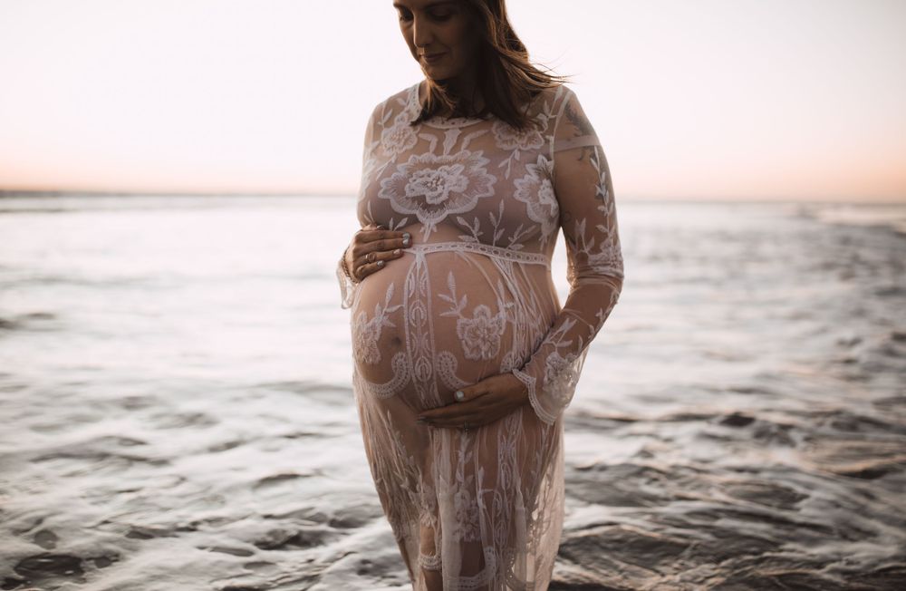 Pregnant woman in lace dress standing in ocean water during sunset.
