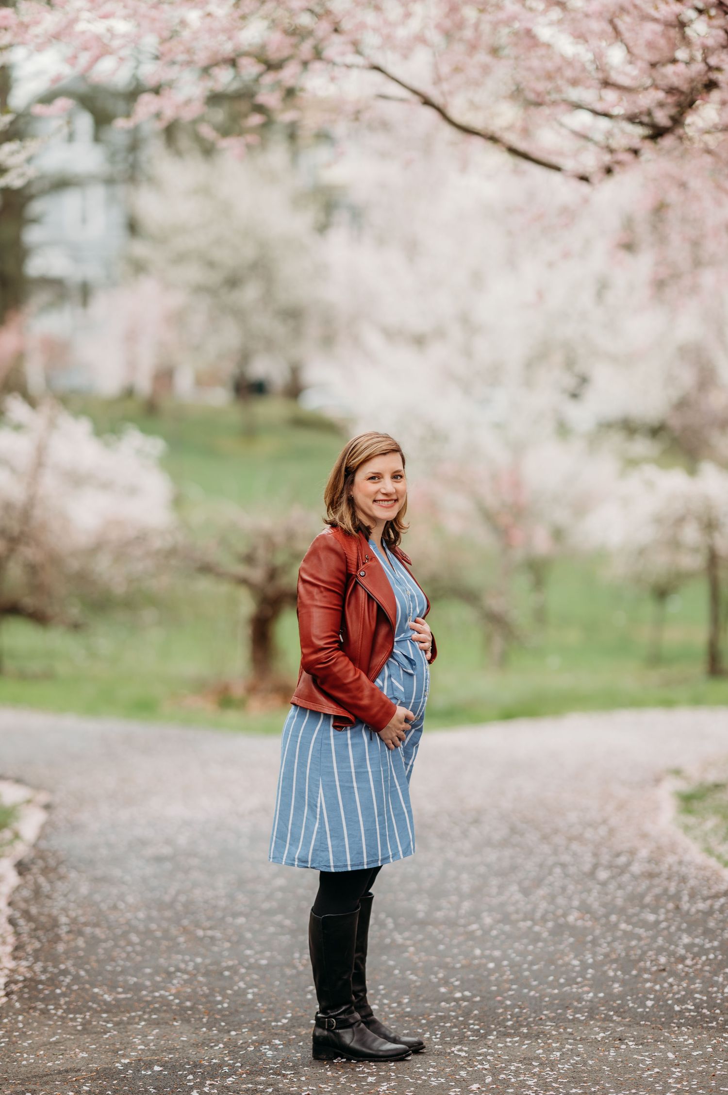 A person in a brown leather jacket and striped dress stands on a path beneath blooming cherry blossoms in spring cradling her baby bump.