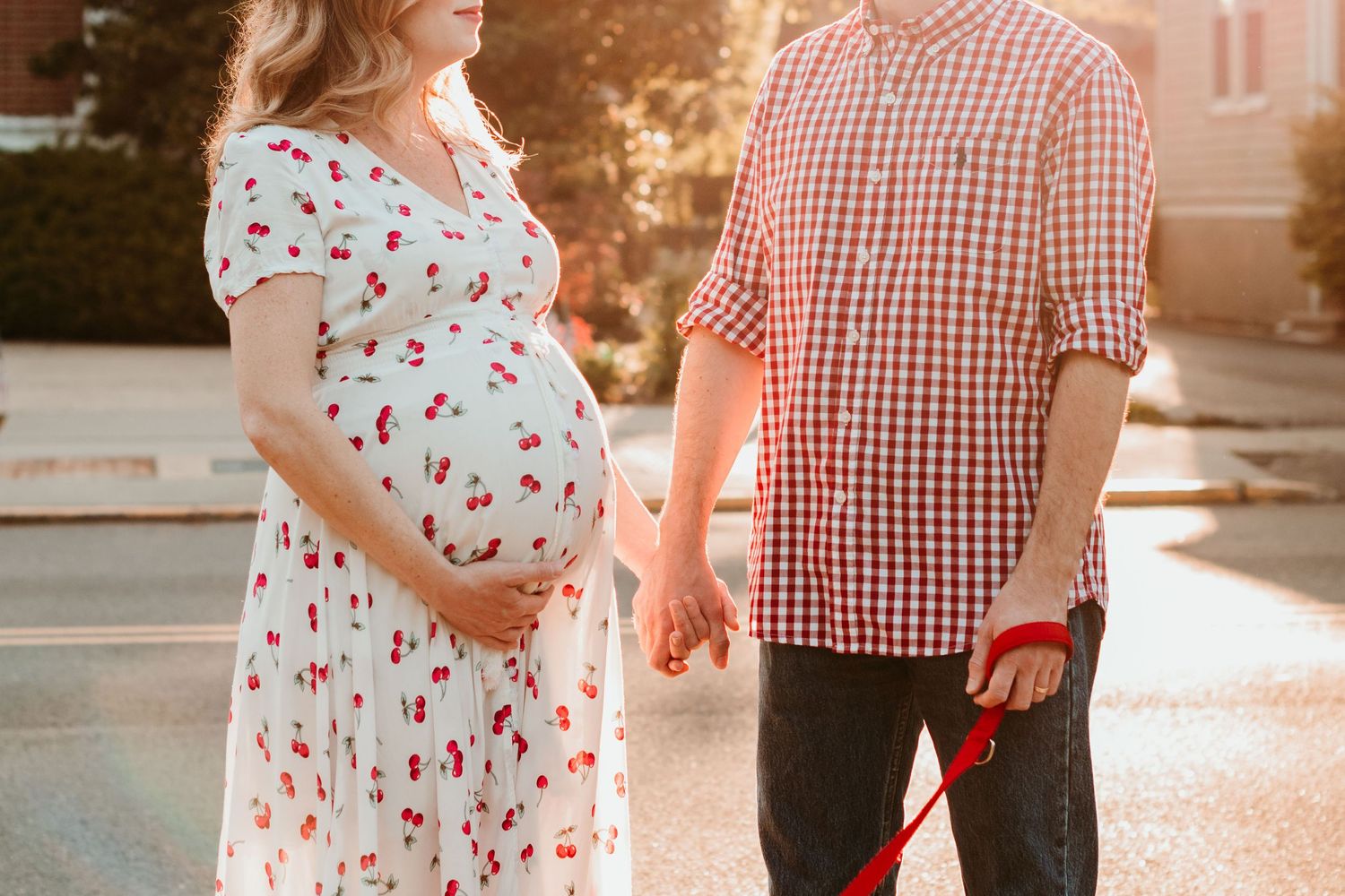 A pregnant person in a floral dress holds hands with their partner wearing a red gingham shirt at sunset in front of the Bloomfield Lirbrary