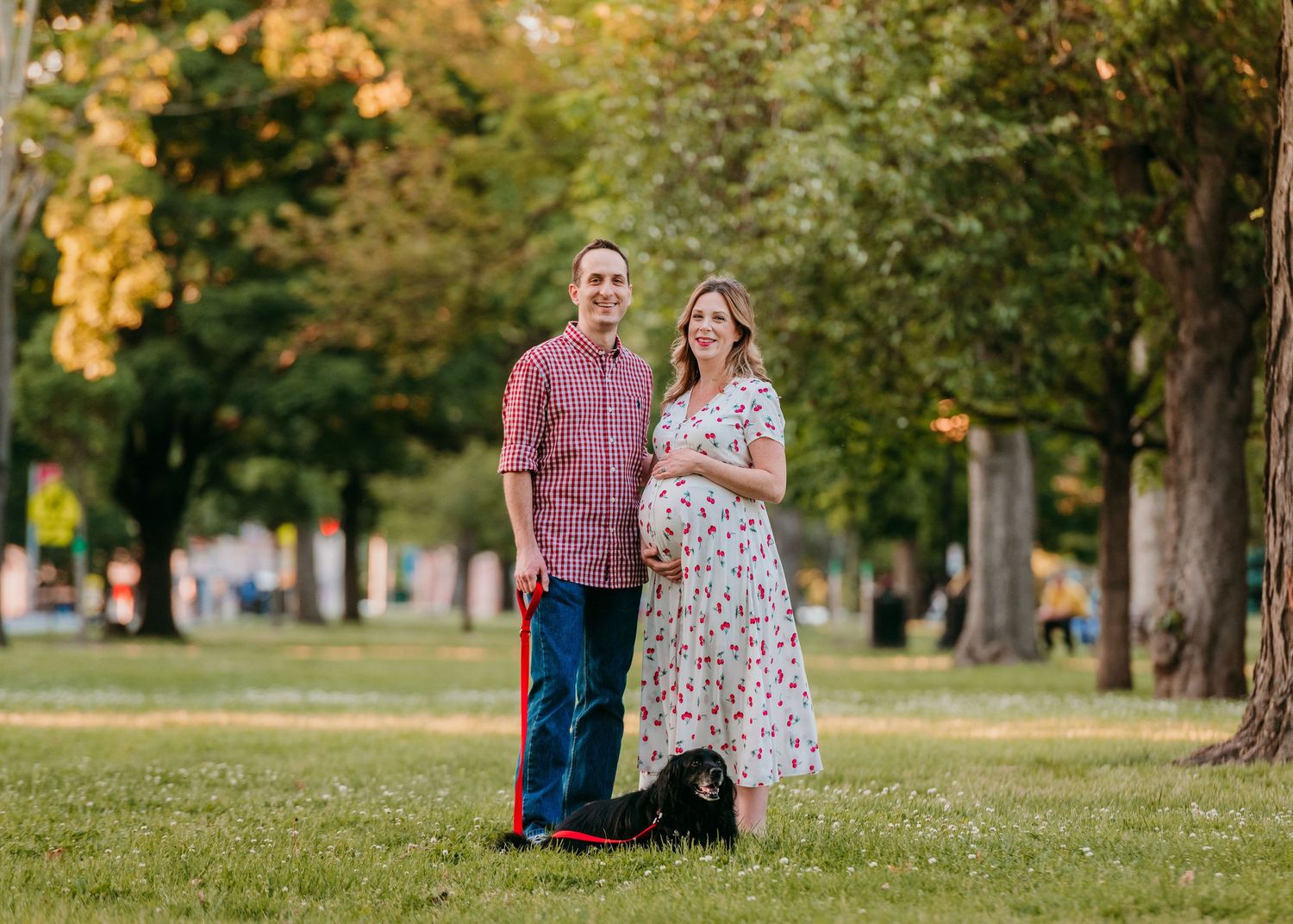 A couple stands together on a grassy lawn with their black dog during a warm autumn evening in the park at Bloomfield green.