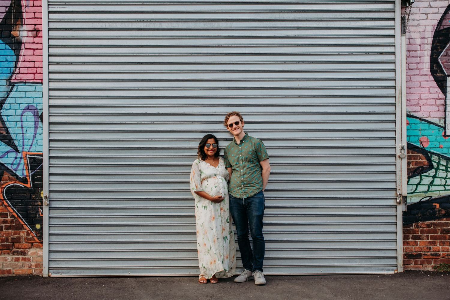 A couple stands together against a gray metal garage door with street art visible on brick walls in Jersey City.