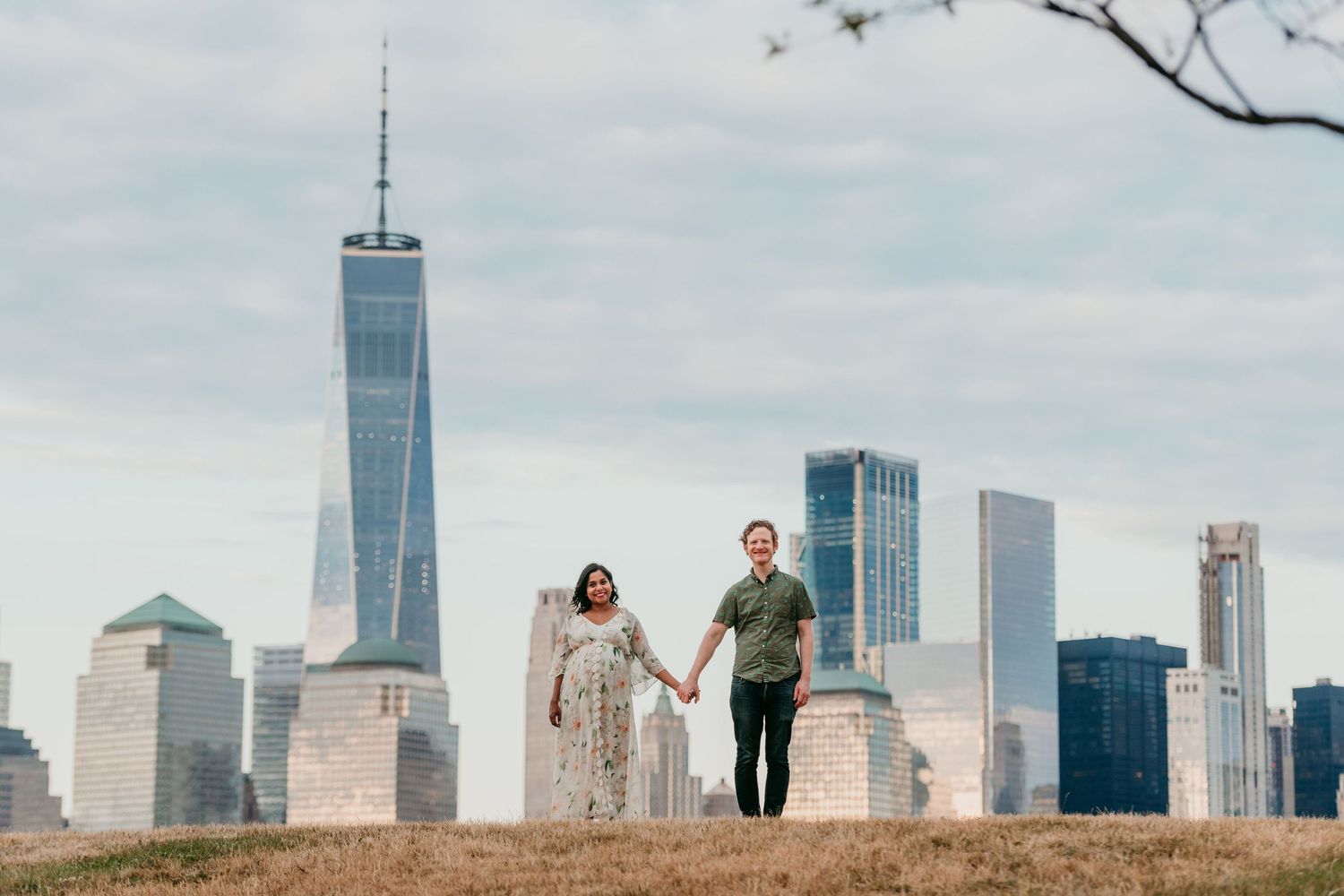 A couple walks hand in hand on a hilltop with the iconic New York City skyline and One World Trade Center in the background in Jersey City.