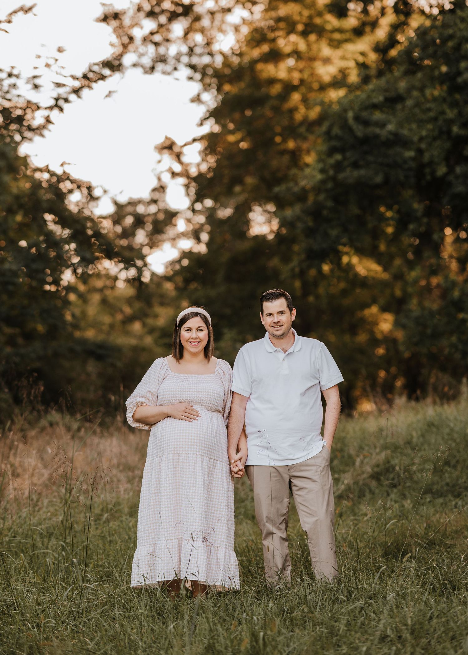 A couple stands together in a golden sunset field, with the expectant mother wearing a flowing white dress in Morris County.