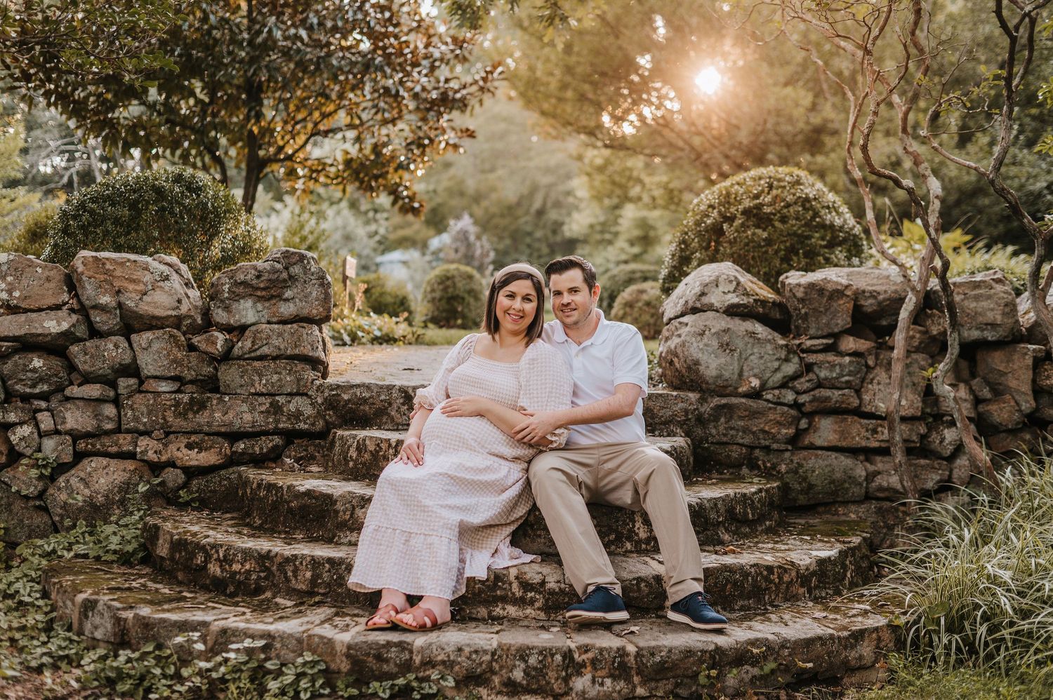 A couple in white clothing sits on rustic stone steps surrounded by lush greenery during golden hour sunset at Cross Estate Gardens.