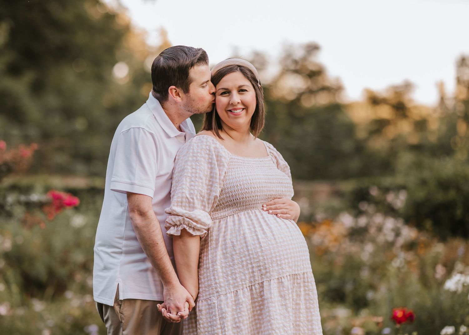 A couple holds hands in a garden setting at sunset while wearing light colored casual clothing to celebrate a pregnancy in Bernardsville.