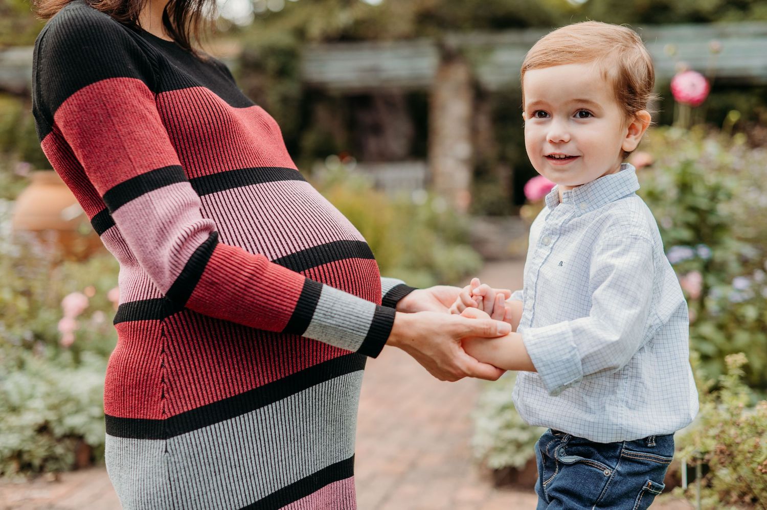 A child in a light-colored shirt holds hands with someone wearing a striped sweater in a garden setting during a maternity session in Morris