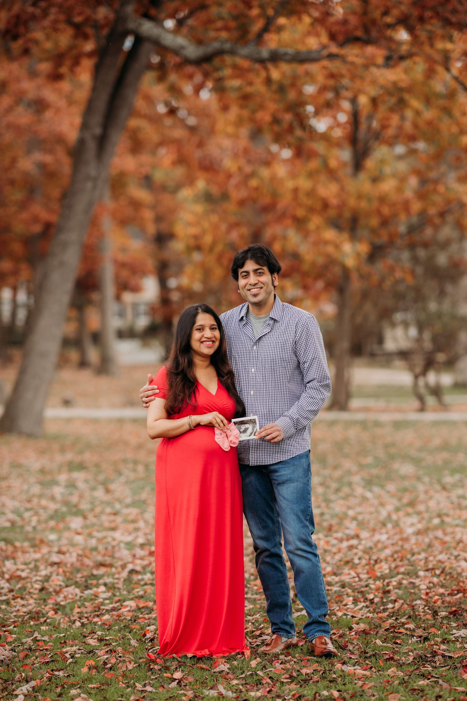 A couple stands together on fallen autumn leaves, with vibrant orange foliage in the background at Verona Park in Essex County.