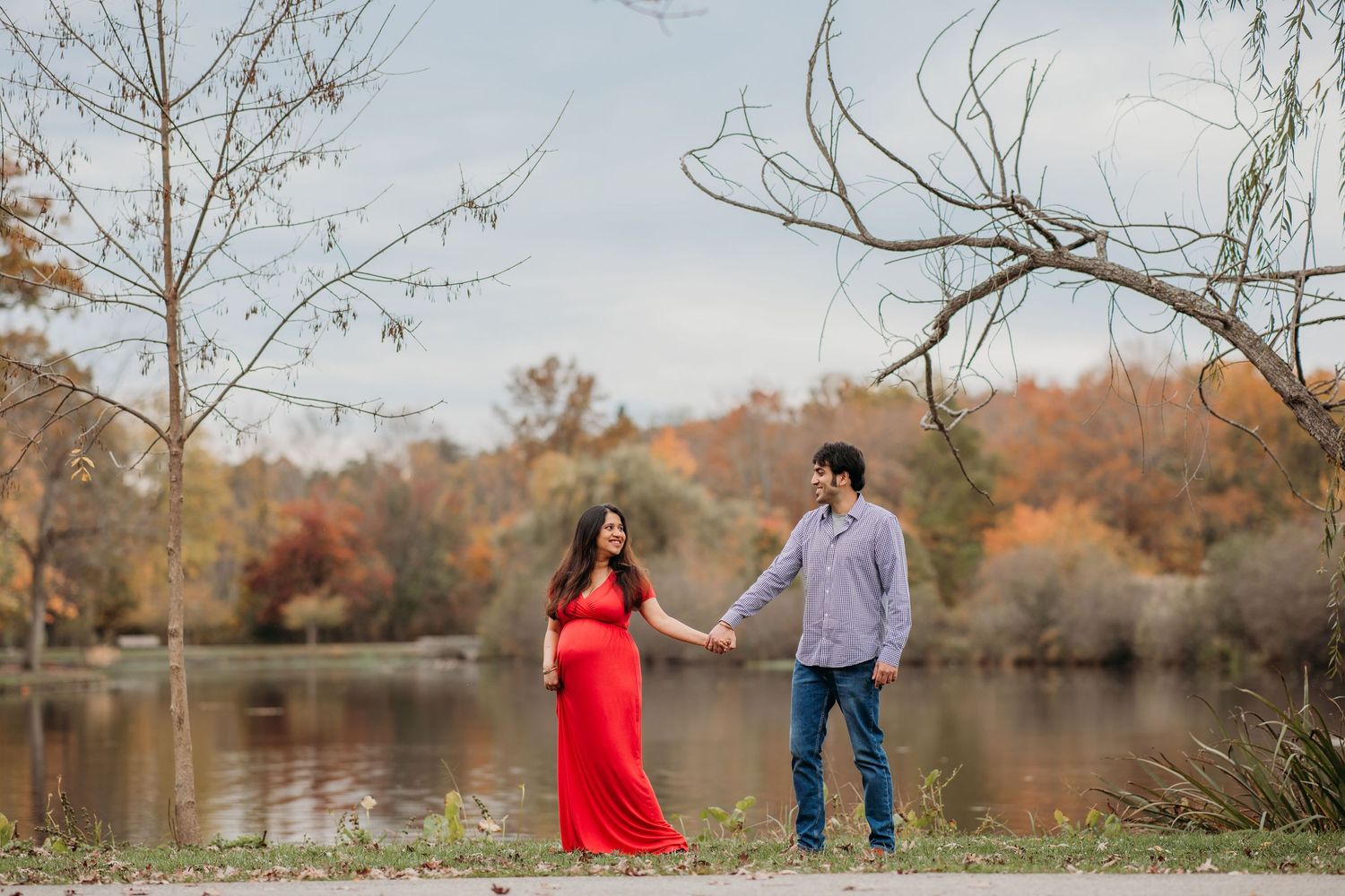 A couple in autumn attire stands by a serene lake surrounded by fall foliage and bare trees at Verona Park.