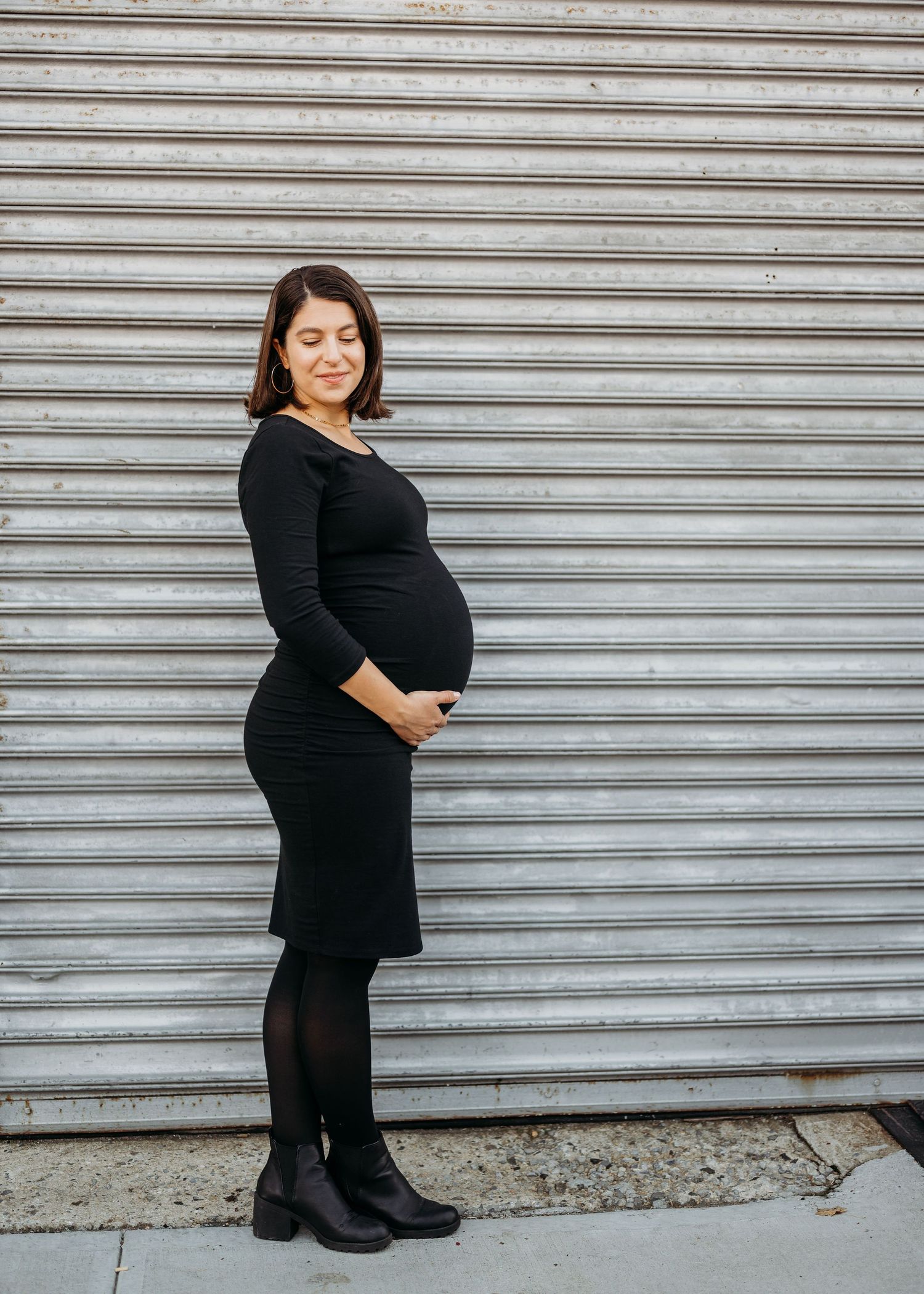 A pregnant woman in a black dress and boots stands against an industrial metal door background in Brooklyn.