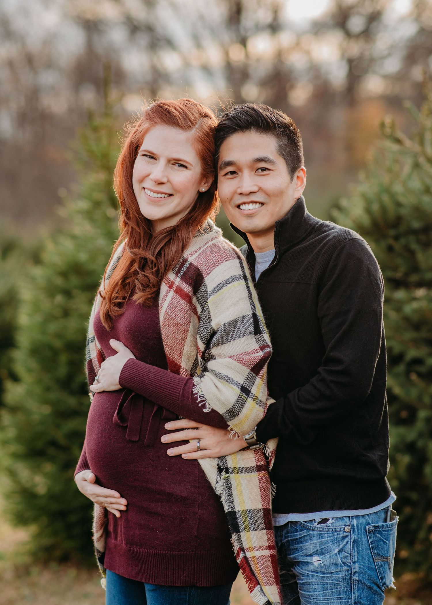 Expectant couple in fall maternity photo session standing among evergreen trees at sunset in Mendham.