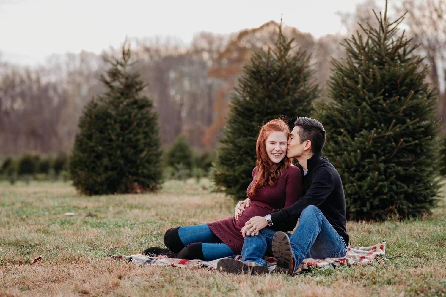 A couple sits on a blanket among evergreen trees at Hidden Pond Christmas tree farm on an autumn day.