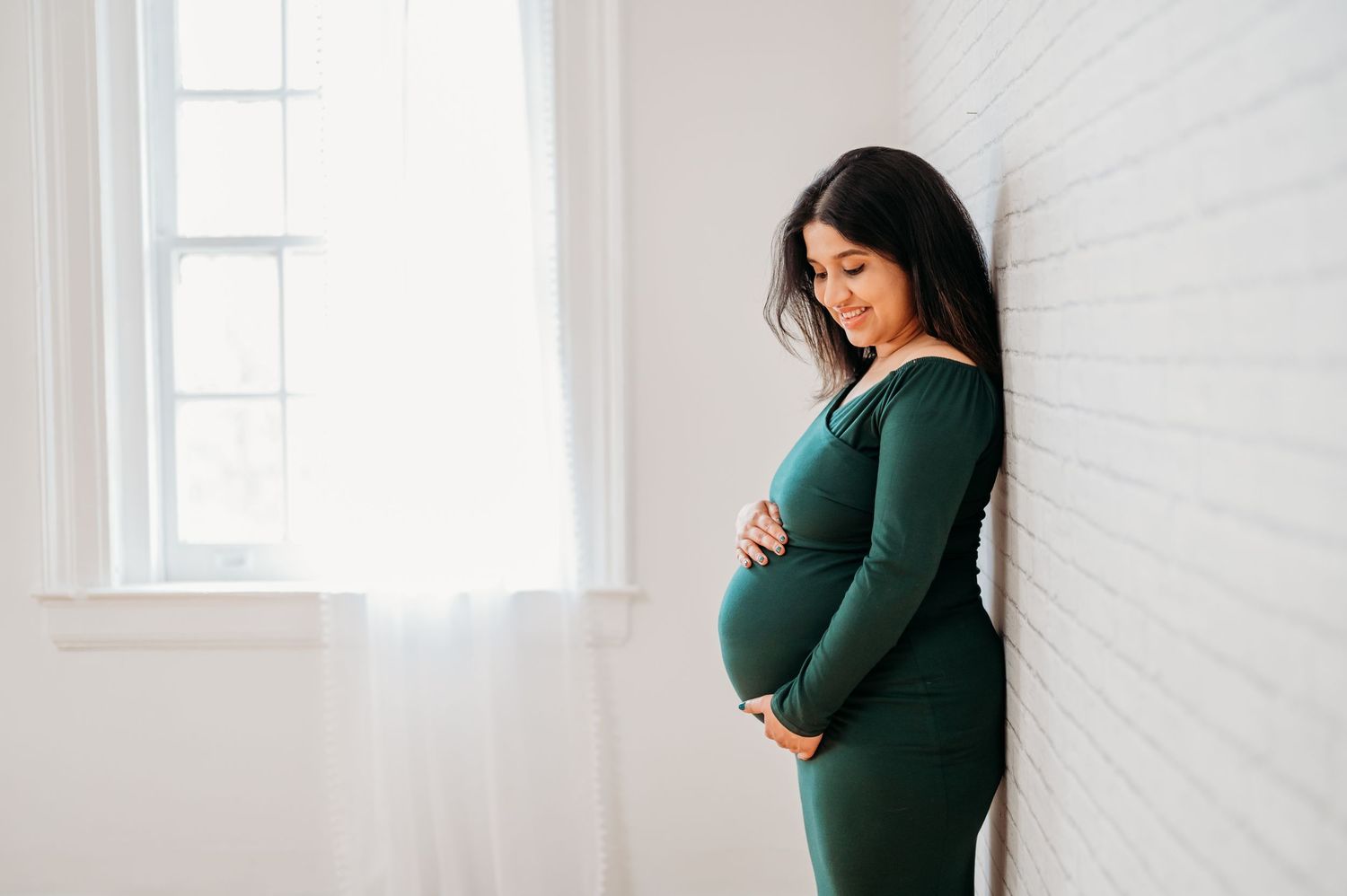 A pregnant woman in a dark green sweater stands by a bright window while cradling her baby bump.