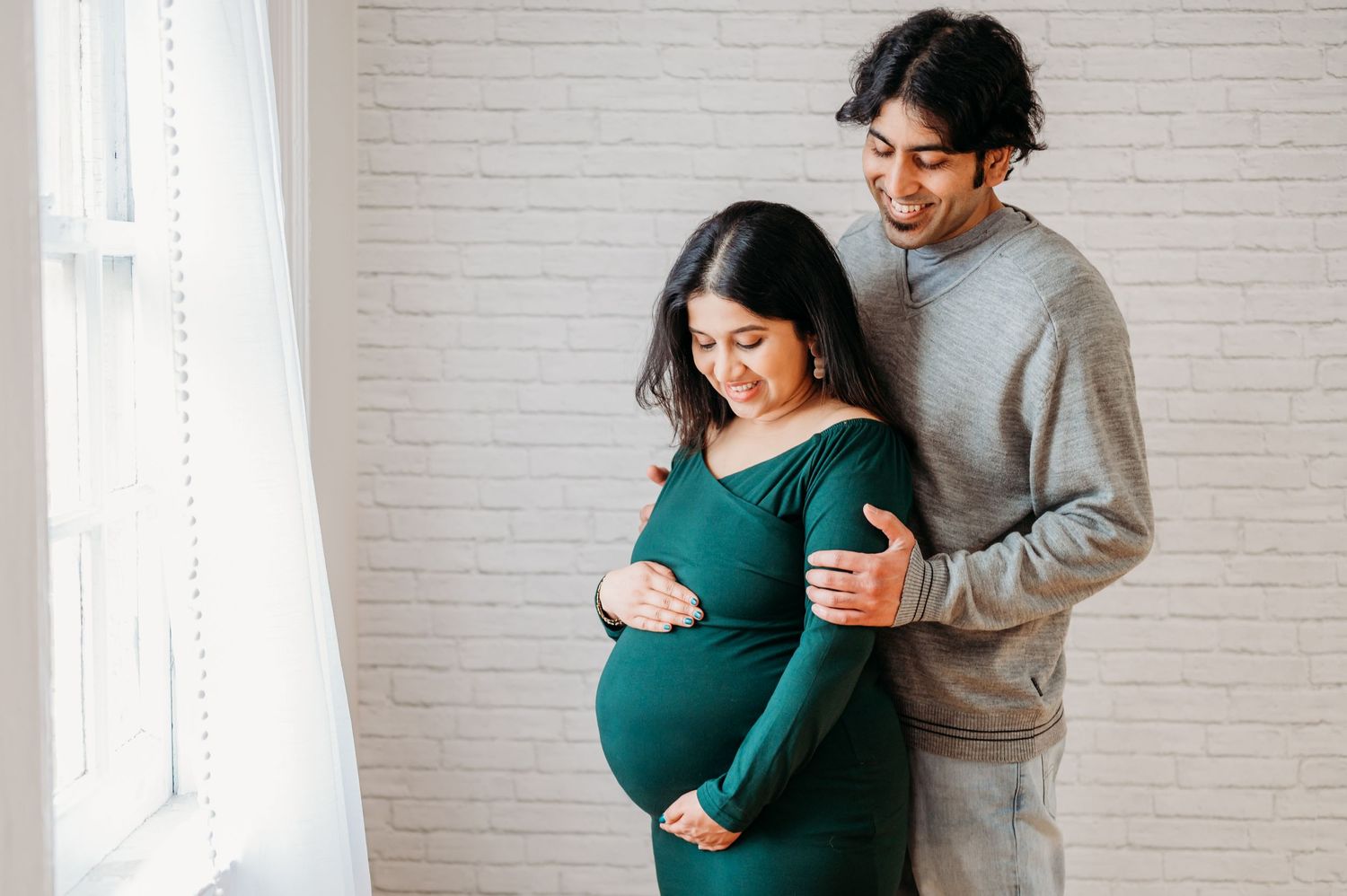 Expectant couple sharing tender moment by window as they lovingly embrace pregnant belly in sunlit room.