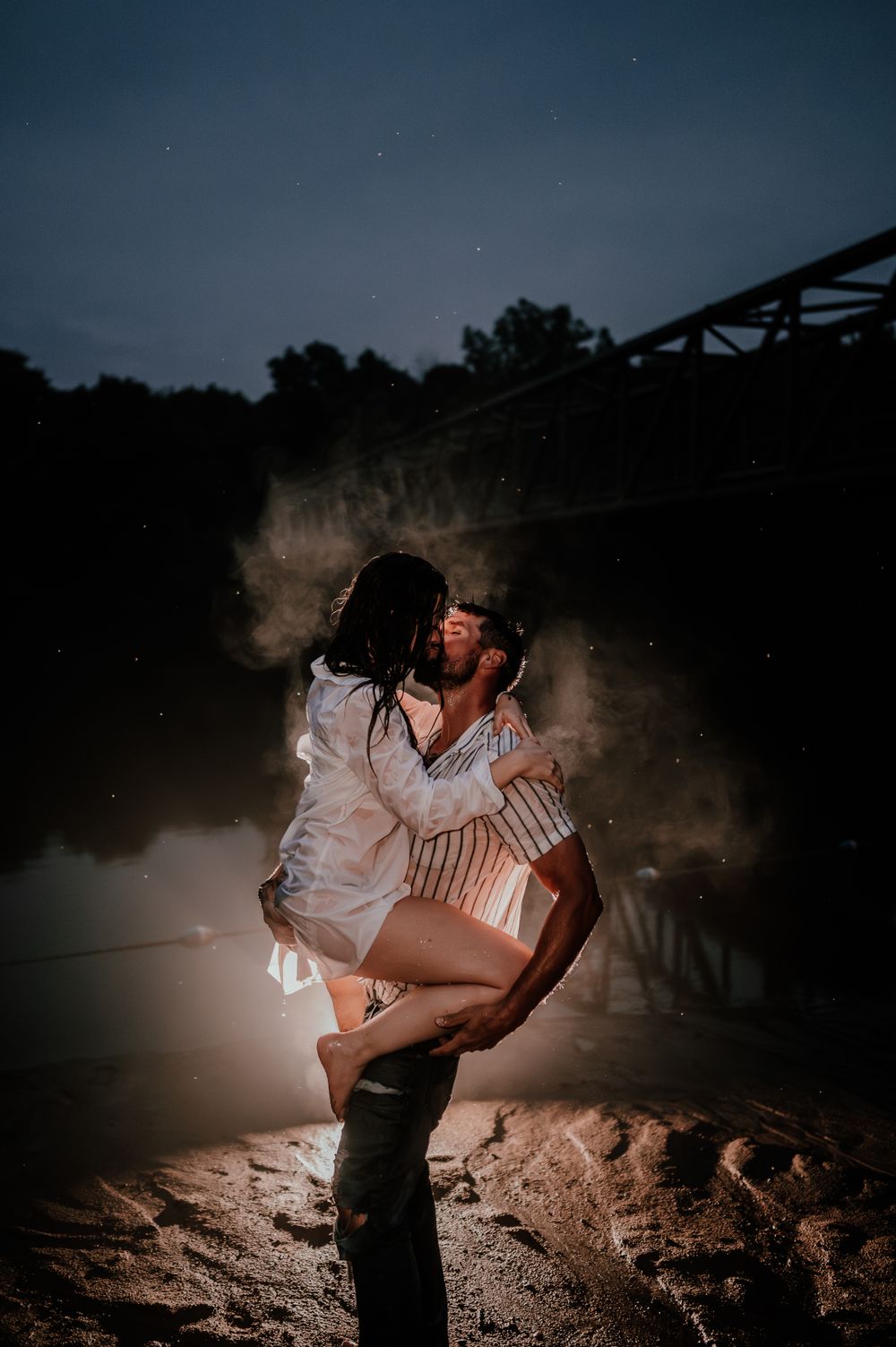 A dramatic nighttime couple's portrait with atmospheric smoke effects and bridge silhouette in background. un couple amoureux sous la pluie