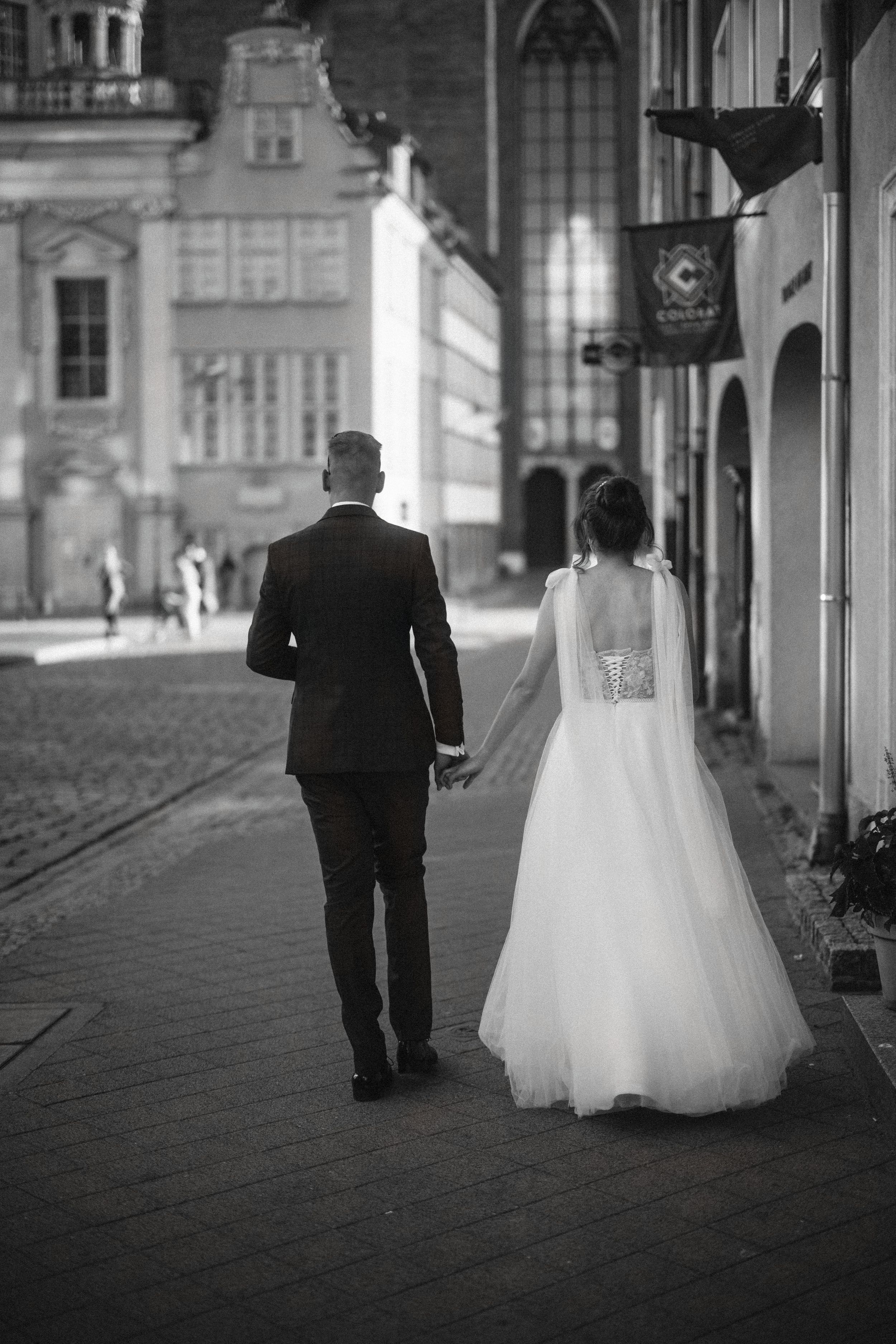 A wedding couple walks hand in hand down a historic cobblestone street in black and white.