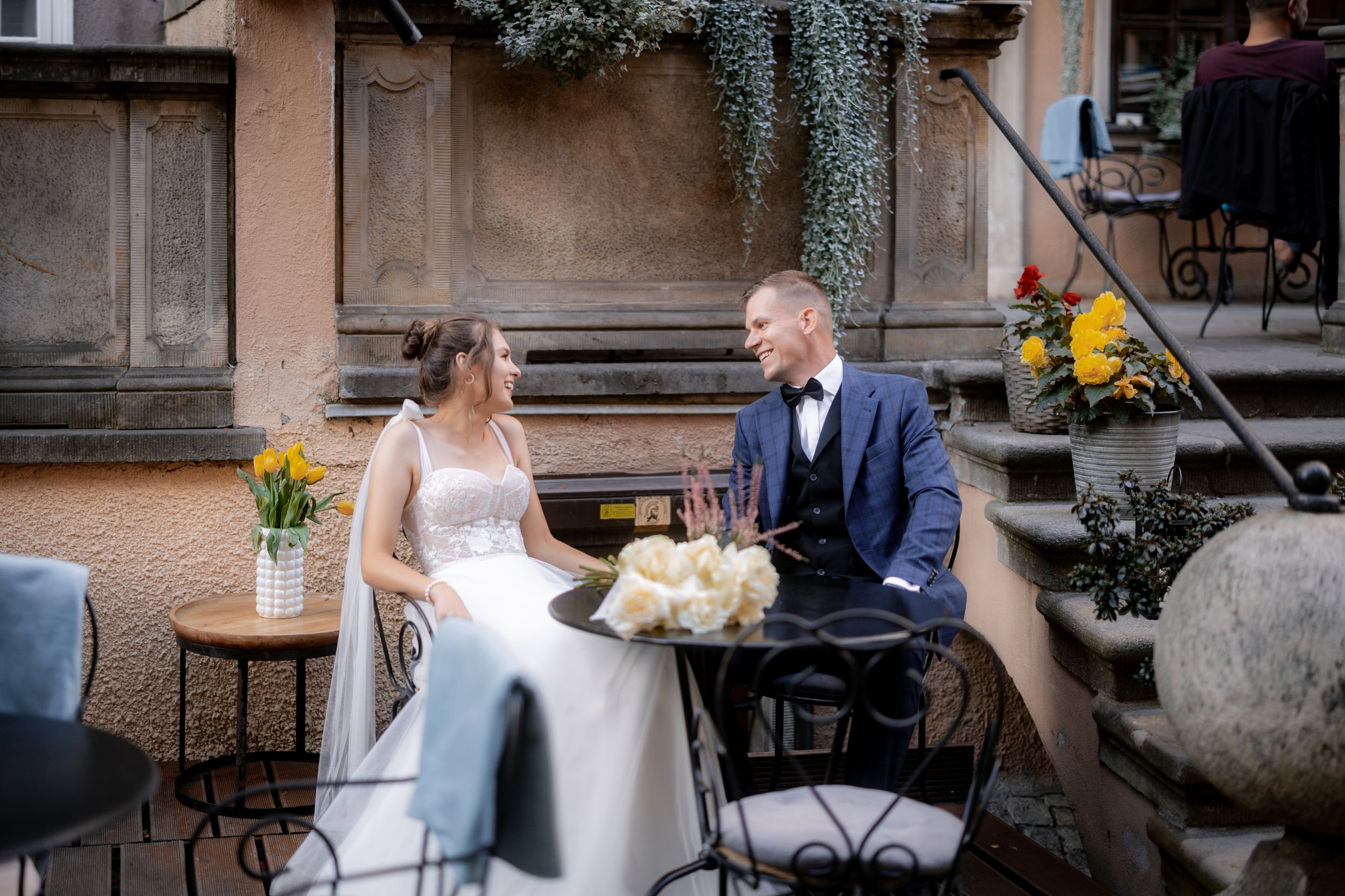 A couple shares a quiet moment at an outdoor cafe table during their wedding celebration.