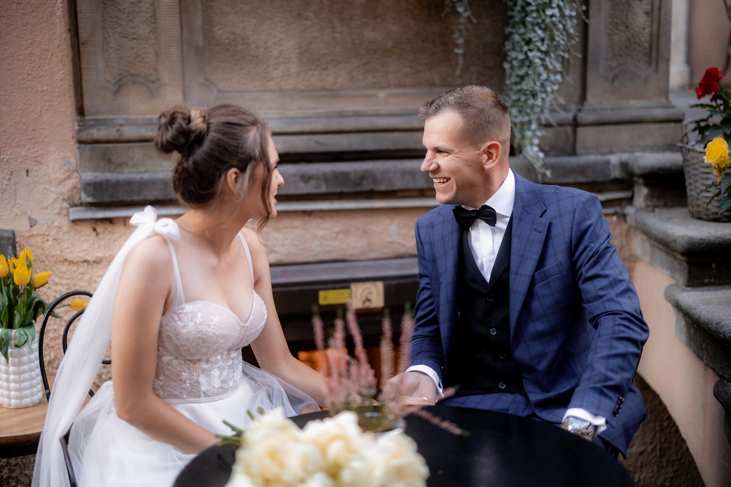 A candid moment between newlyweds seated at an outdoor cafe table with spring flowers.