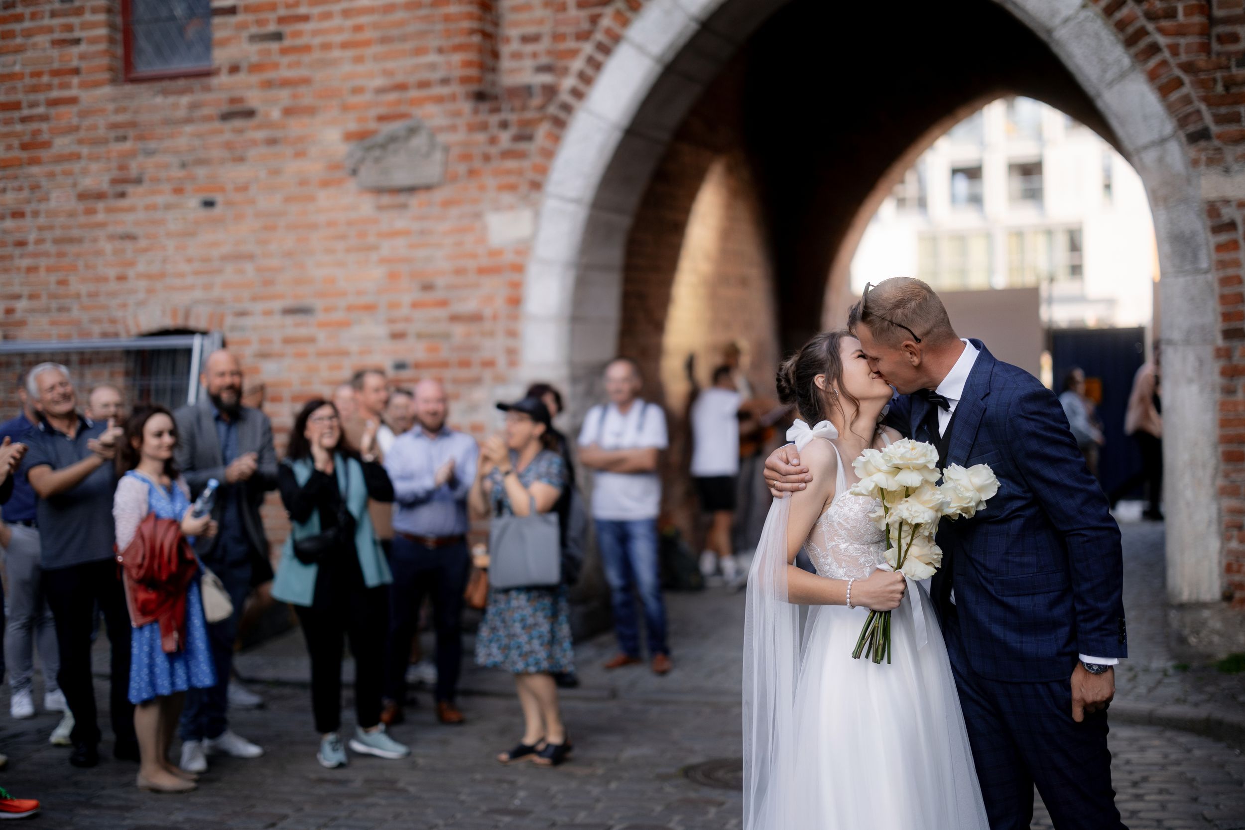 A newlywed couple shares a romantic moment under a brick archway as guests look on during their wedding celebration.