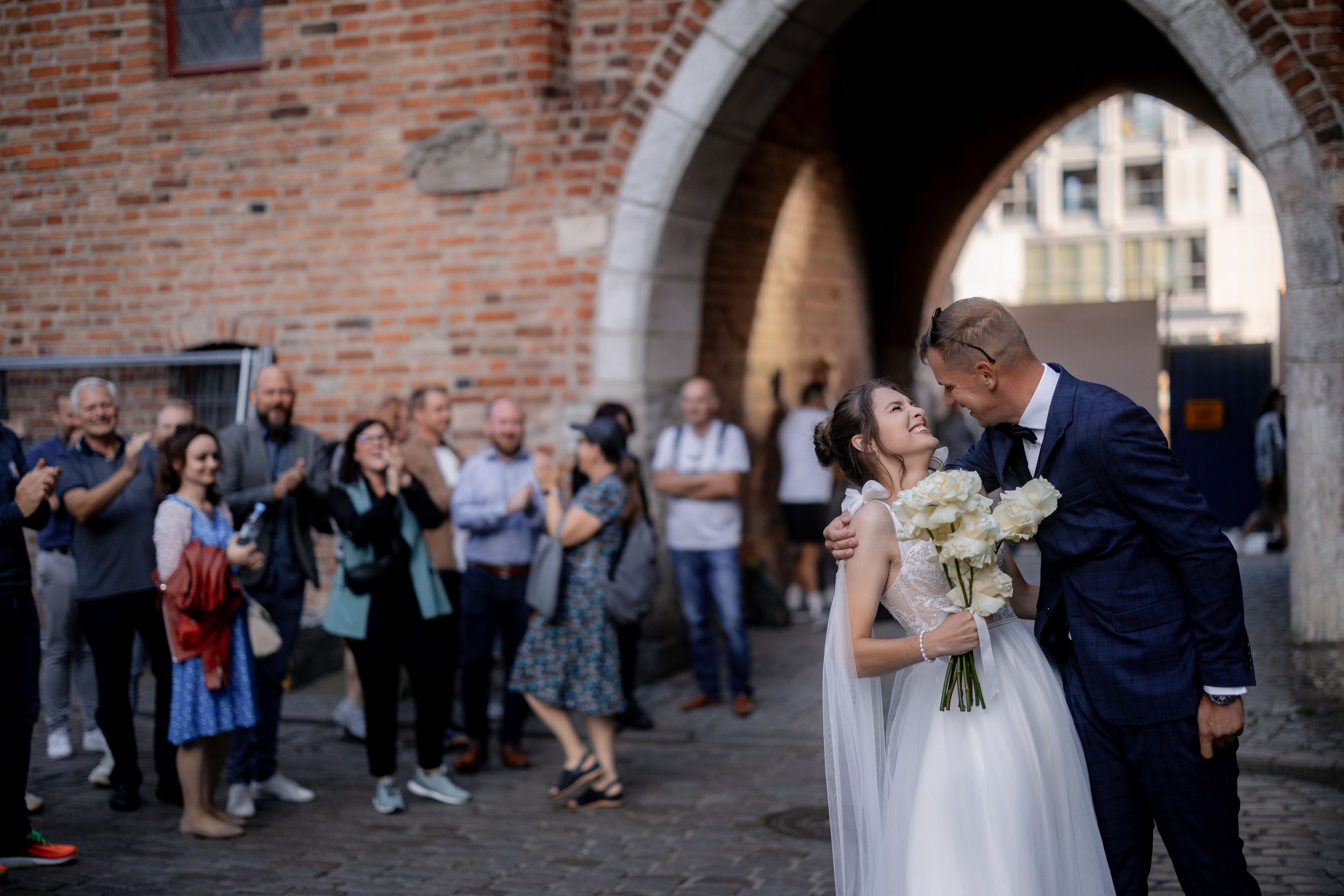 A romantic wedding moment under a brick archway with guests watching and taking photos.