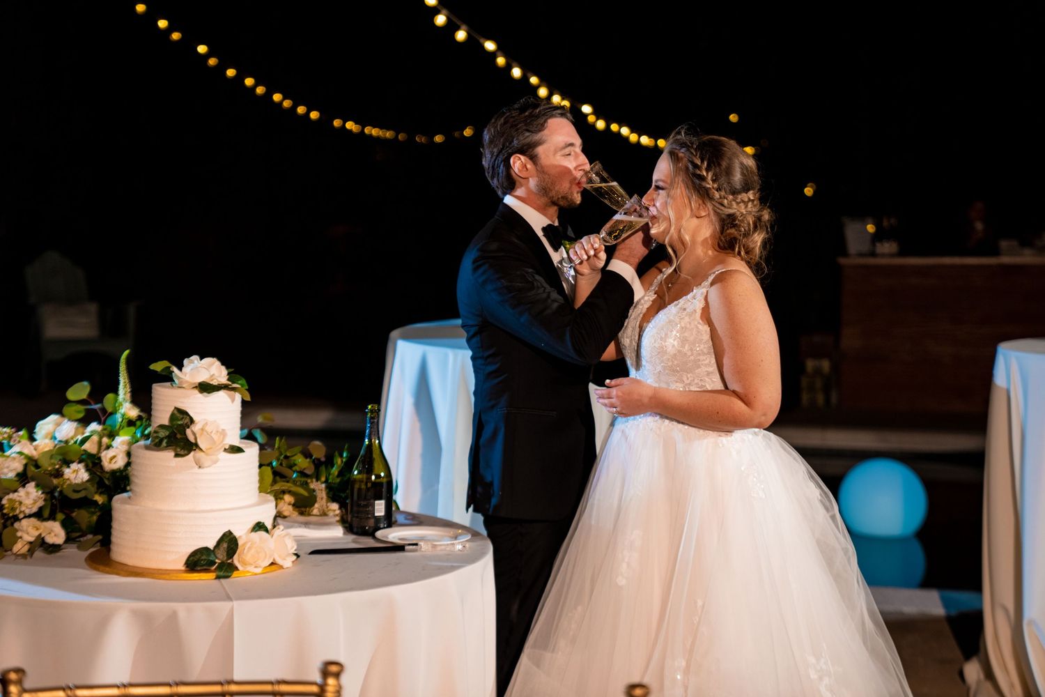 Reception moment captured with wedding cake and string lights in background.