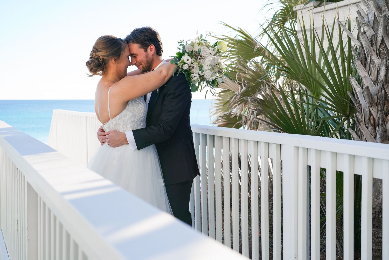 Romantic wedding moment on a white beachfront balcony with palm trees.