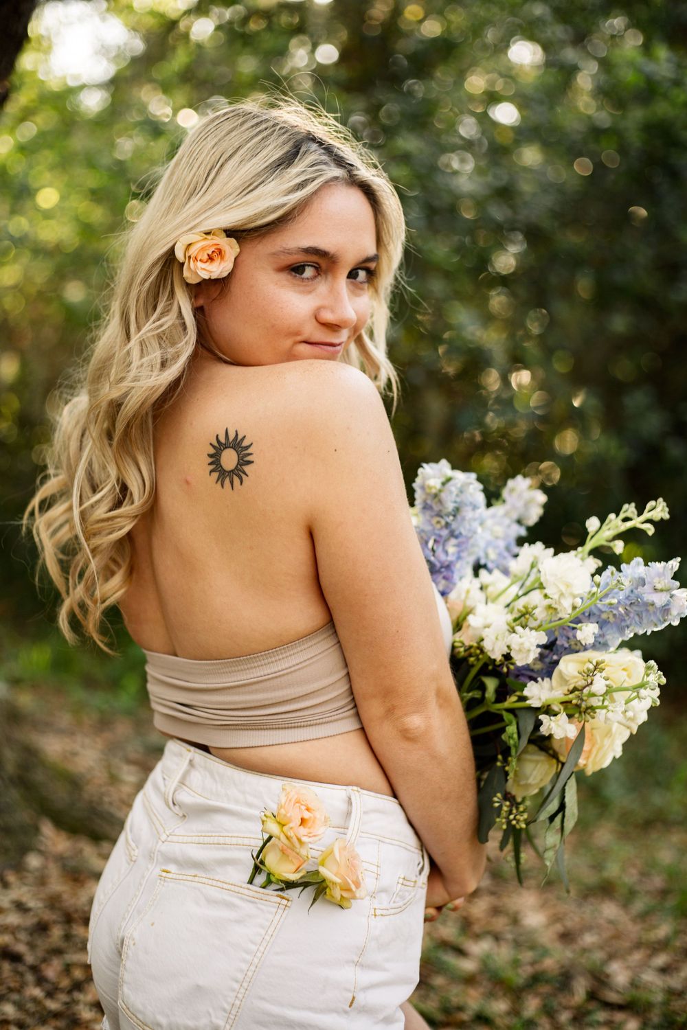 Outdoor portrait showing a sun tattoo detail and floral bouquet against lush greenery.