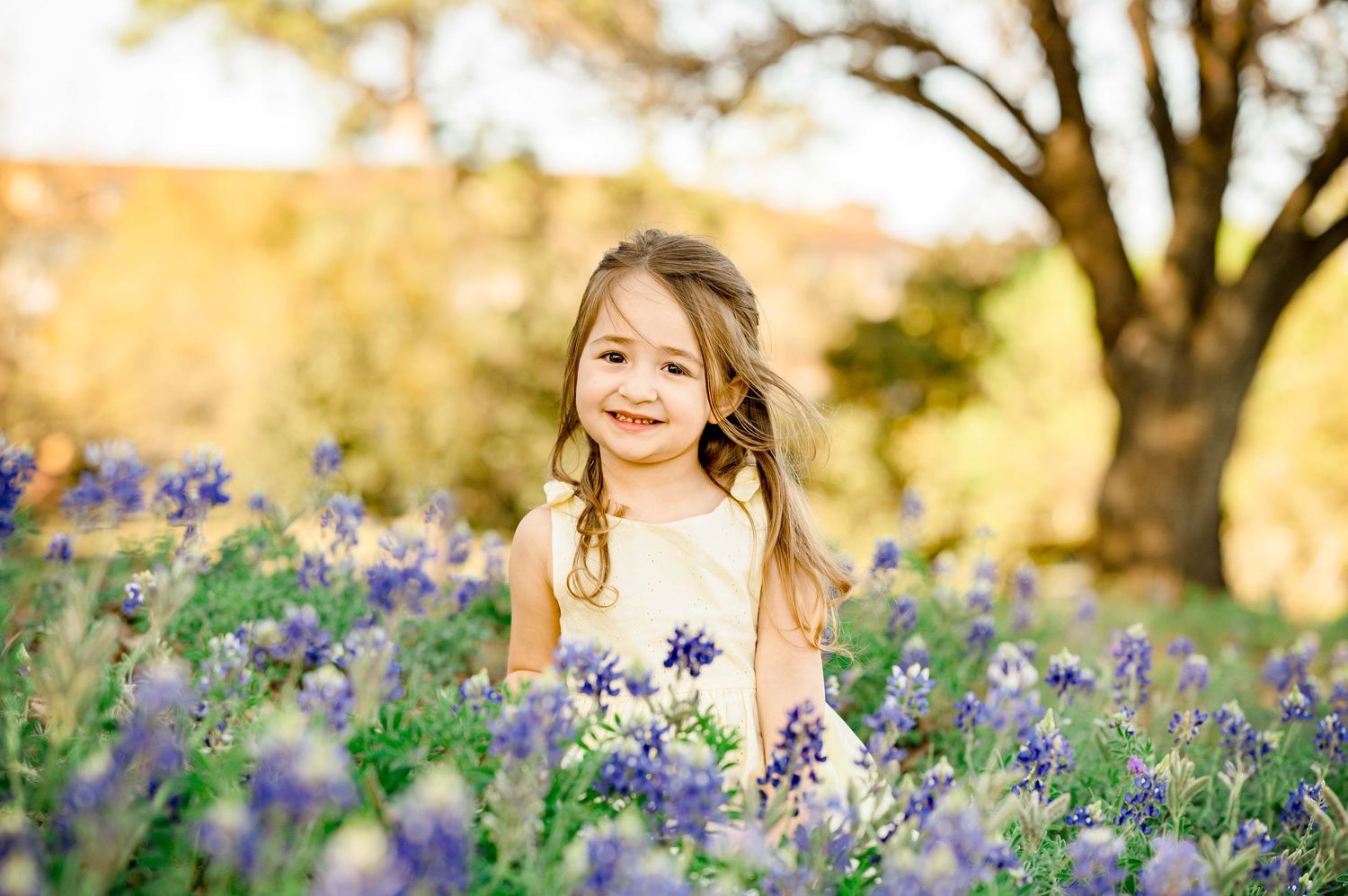 little girl wearing a yellow dress sitting a field of bluebonnets during the golden hour