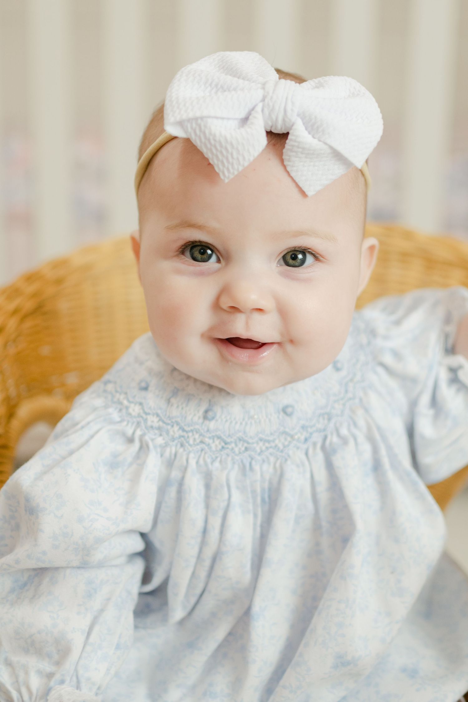 Adorable baby girl in blue smocked dress with white bow headband smiling in a wicker chair.