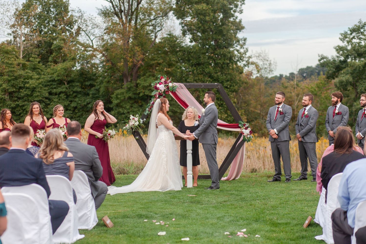 A wedding ceremony takes place outdoors on a grassy lawn with an elegant white and burgundy draped arch.
