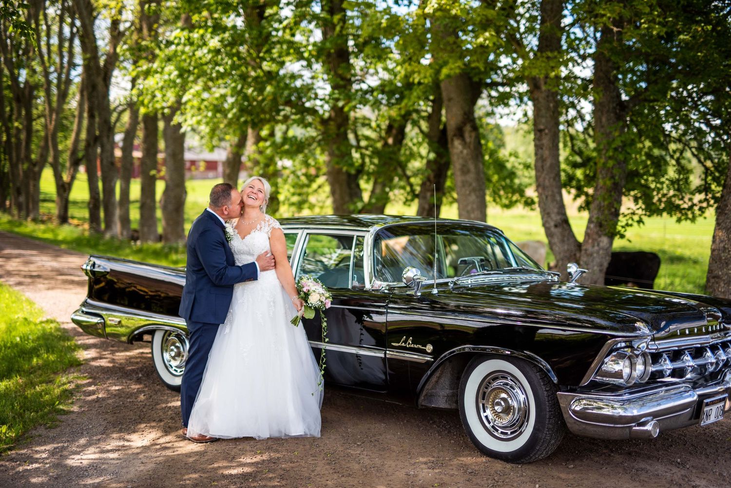 Bride and groom posing beside a classic black vintage car under green trees.