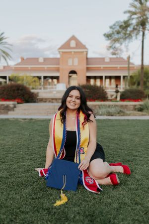 Graduate in colorful stole and red shoes poses playfully on grass with brick building in background.