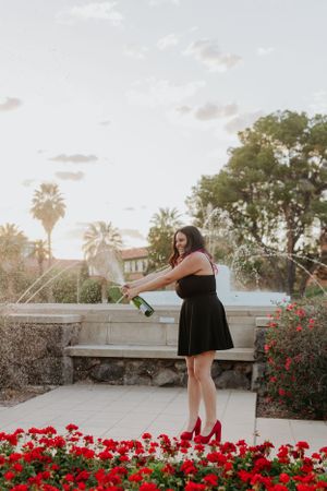 Graduate in black dress celebrating near red flower garden during sunset.