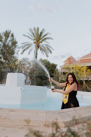 Graduate celebrates by fountain with champagne spray at sunset near palm trees.